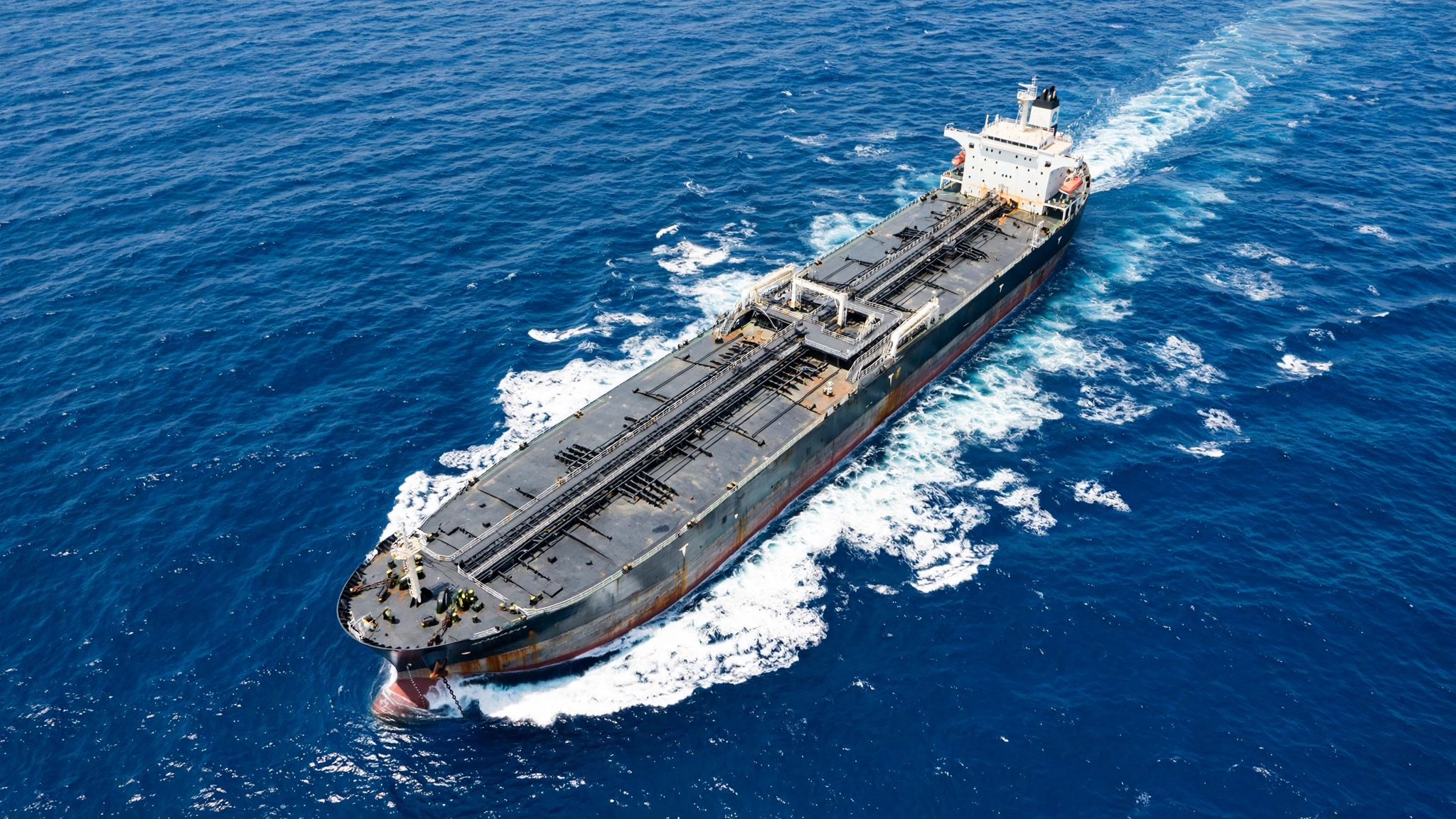 An oil tanker sailing on open blue water with a white wake trailing behind it, seen from above.