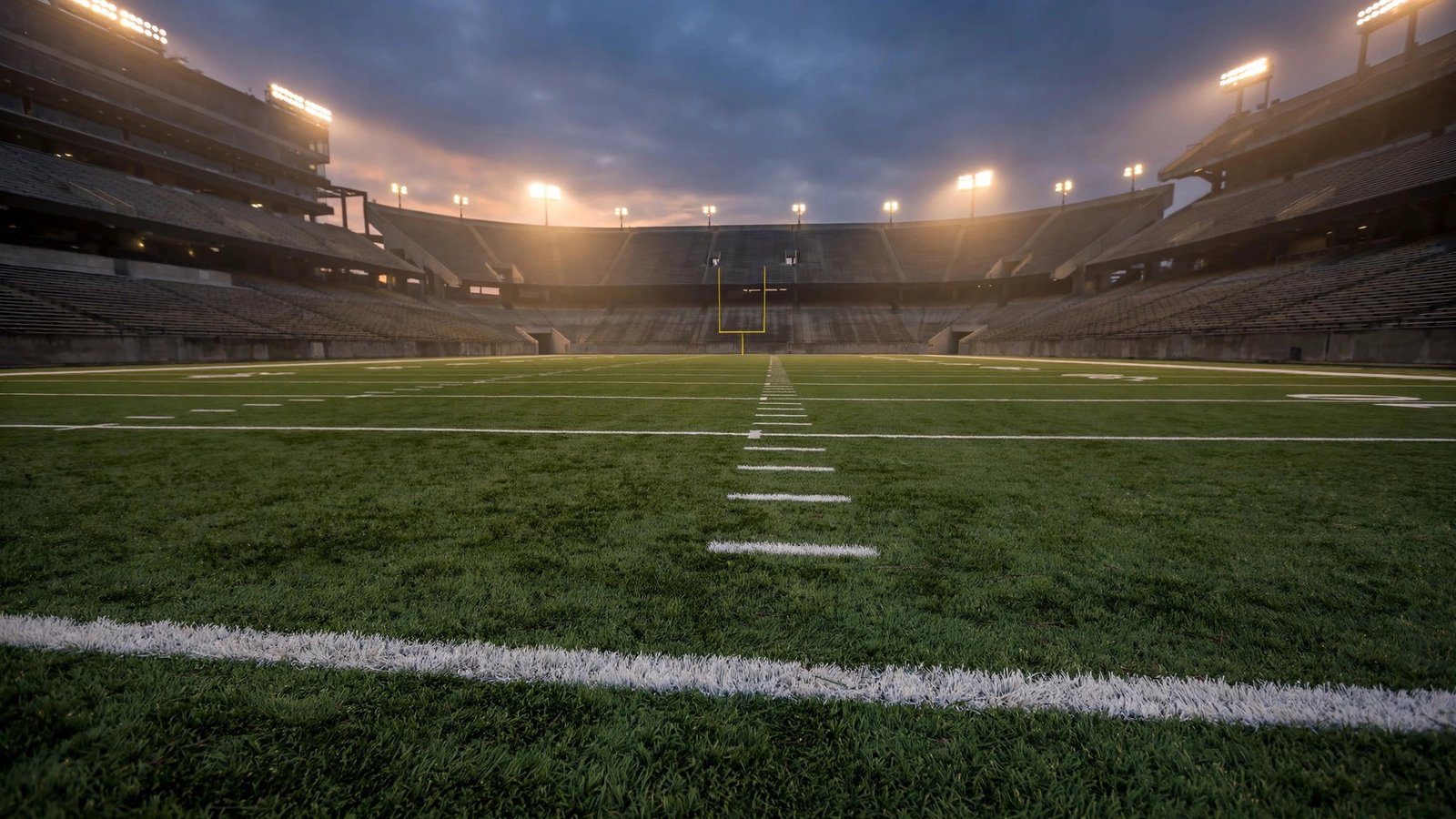An empty football stadium field at dusk, with yard lines stretching toward a goalpost.