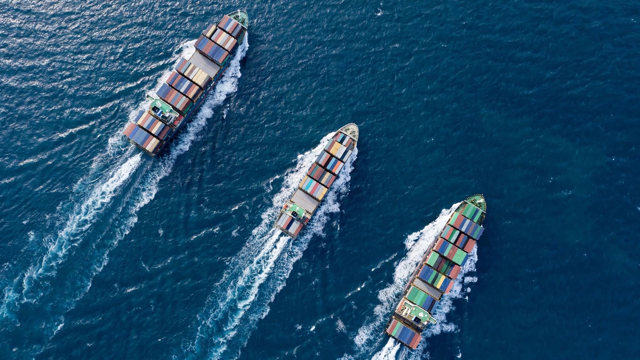 An aerial view of three container cargo ships sailing across open blue water, leaving white wakes.