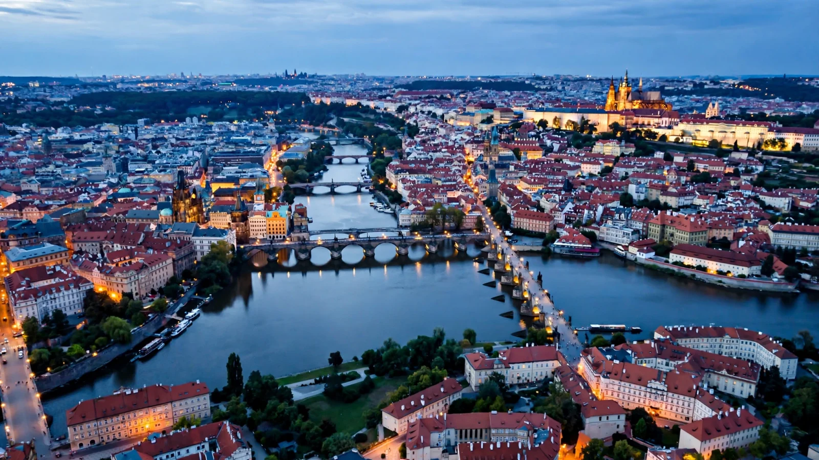 A peaceful aerial view of a European capital city at dusk, red rooftops and church spires along a calm river, city lights just coming on.