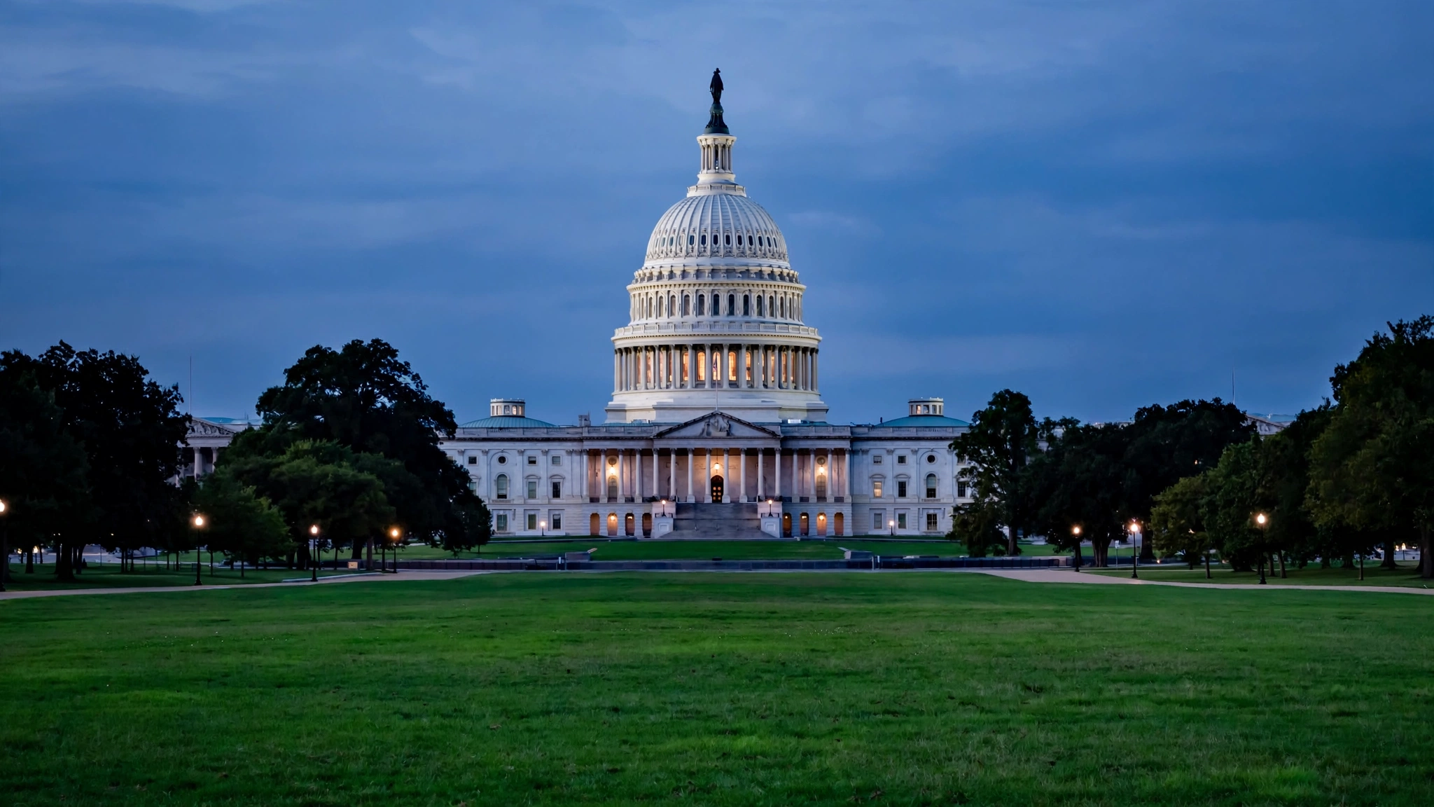 The U.S. Capitol dome illuminated at dusk, viewed from across a wide green lawn lined with trees and lampposts, no people in sight.