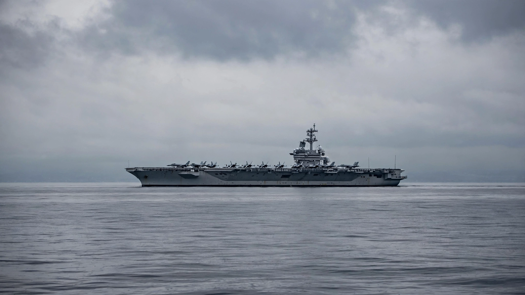 An aircraft carrier with fighter jets on its flight deck sailing on calm water under an overcast sky