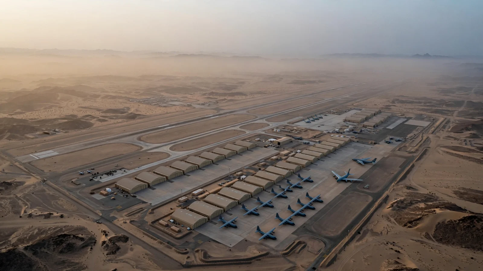 Aerial view of a military airbase in a desert at dawn, with runways, hangars and parked transport aircraft.