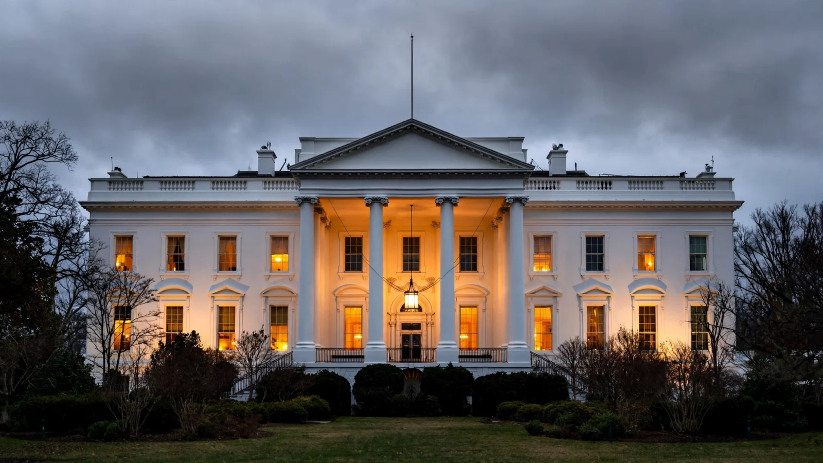 The White House exterior at dusk, with lights on in the windows under a cloudy sky.