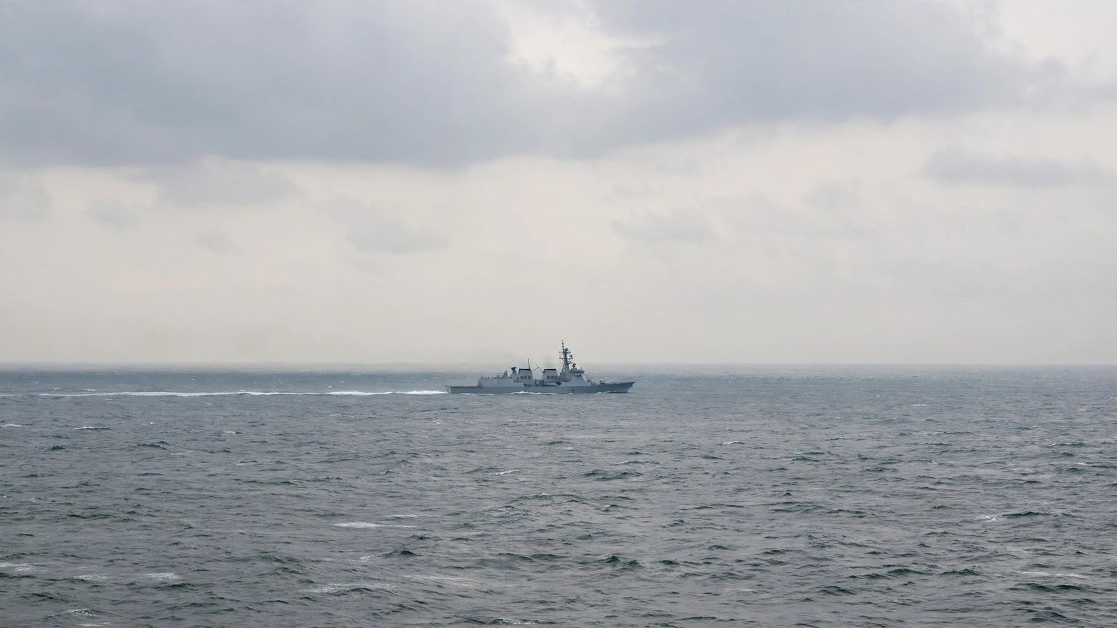 A grey naval warship sailing alone on open sea under a cloudy sky