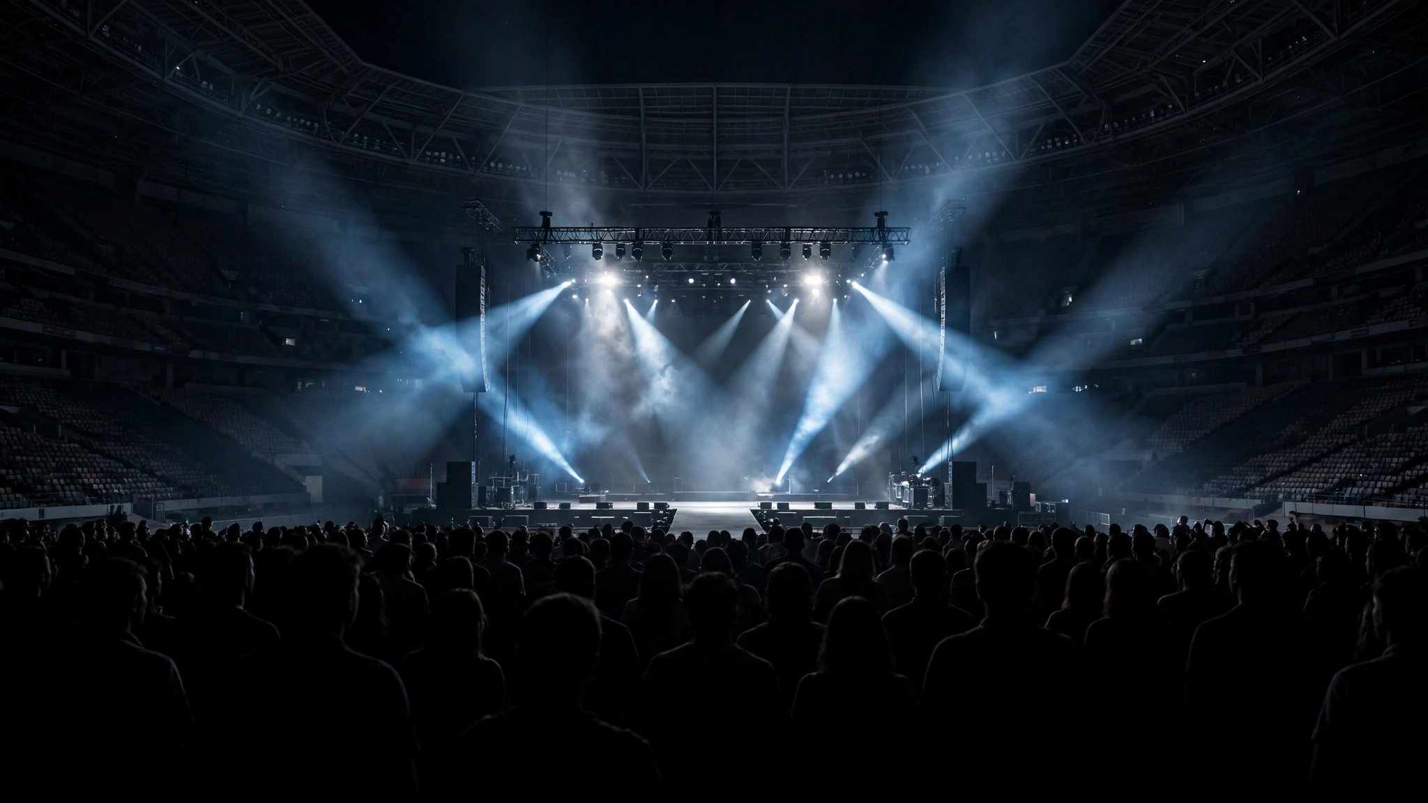 An empty concert stage inside a large arena lit by spotlights, with a silhouetted crowd in the foreground
