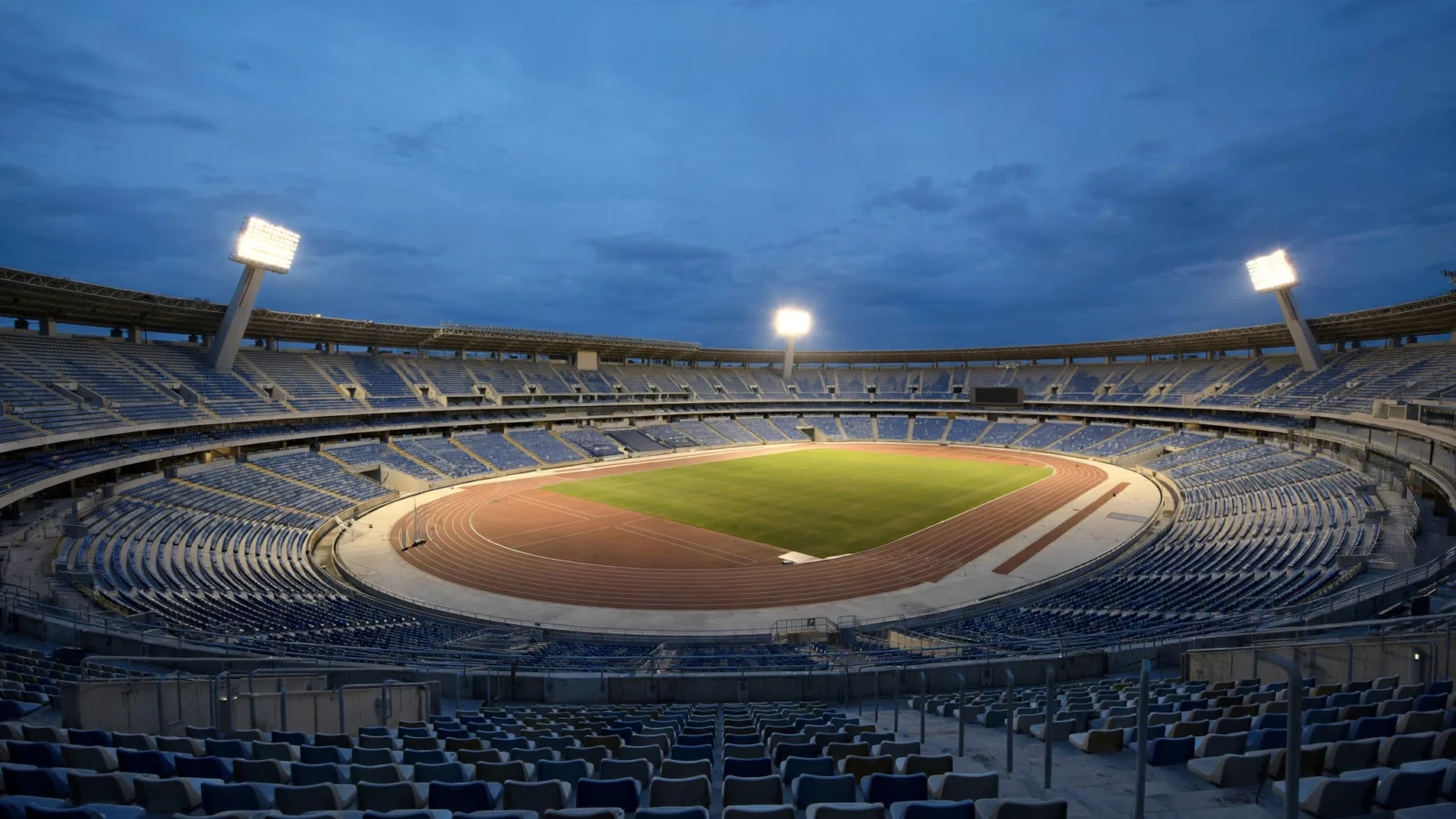 An empty outdoor stadium with a running track and grass field lit by floodlights at dusk