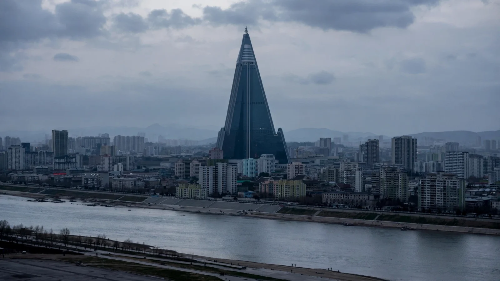 Illustrative photo of the Pyongyang skyline at dusk, with a tall pyramid-shaped hotel tower and apartment blocks along a river.