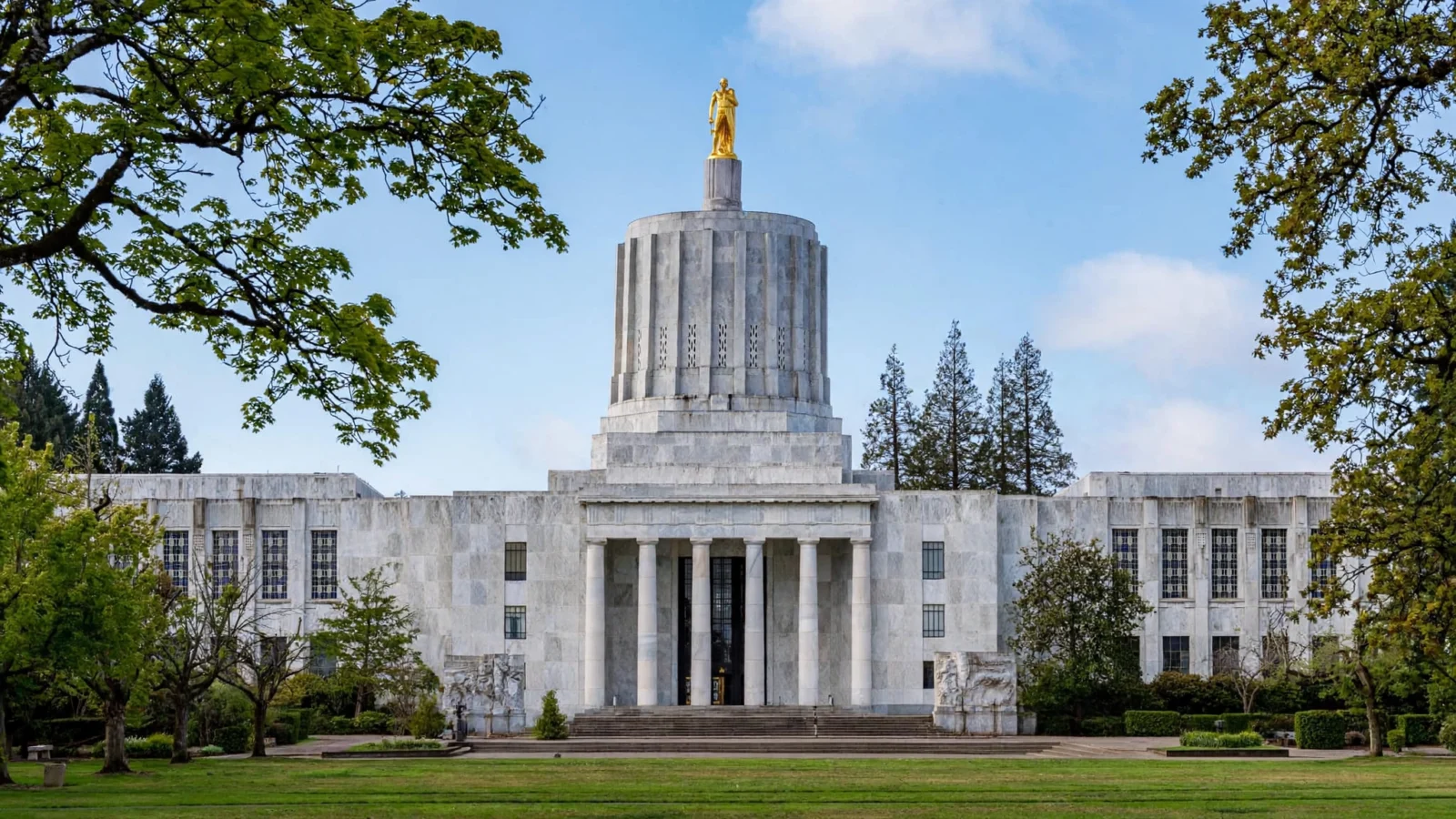 The Oregon State Capitol in Salem, a white marble building with a cylindrical rotunda topped by a gilded statue, front colonnade, green lawn and trees under a blue sky; no people.
