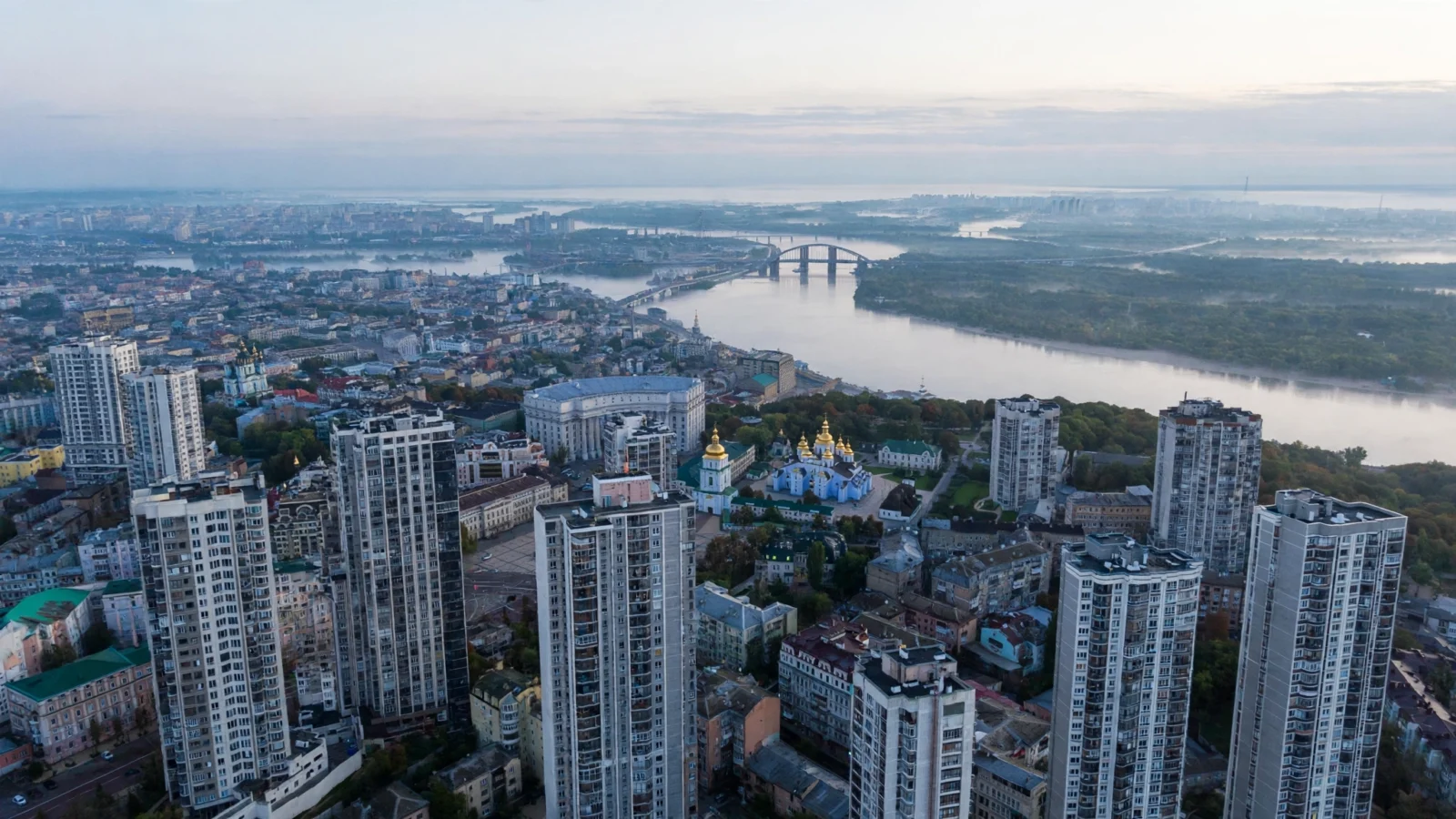 Aerial view of the Kyiv city skyline at dawn, with residential high-rise buildings and golden church domes along the Dnipro River