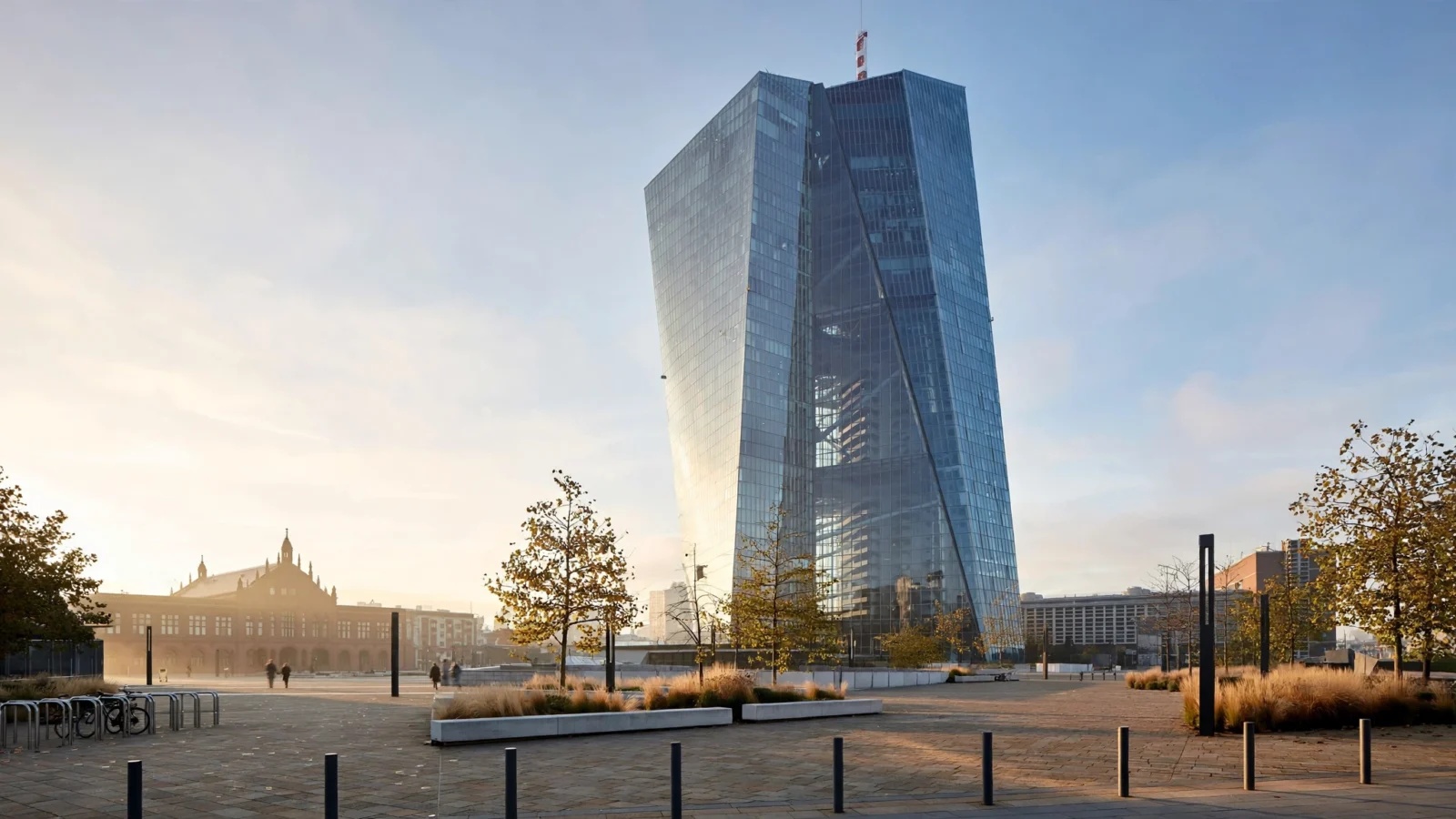 A tall glass office tower in Frankfurt seen from a paved plaza with small trees, bicycle racks and a few distant pedestrians.