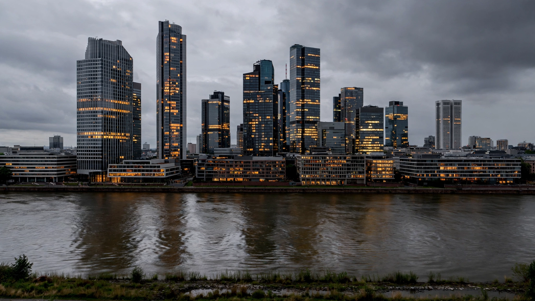 Office towers with lit windows in Frankfurt seen across a wide river under a cloudy evening sky.