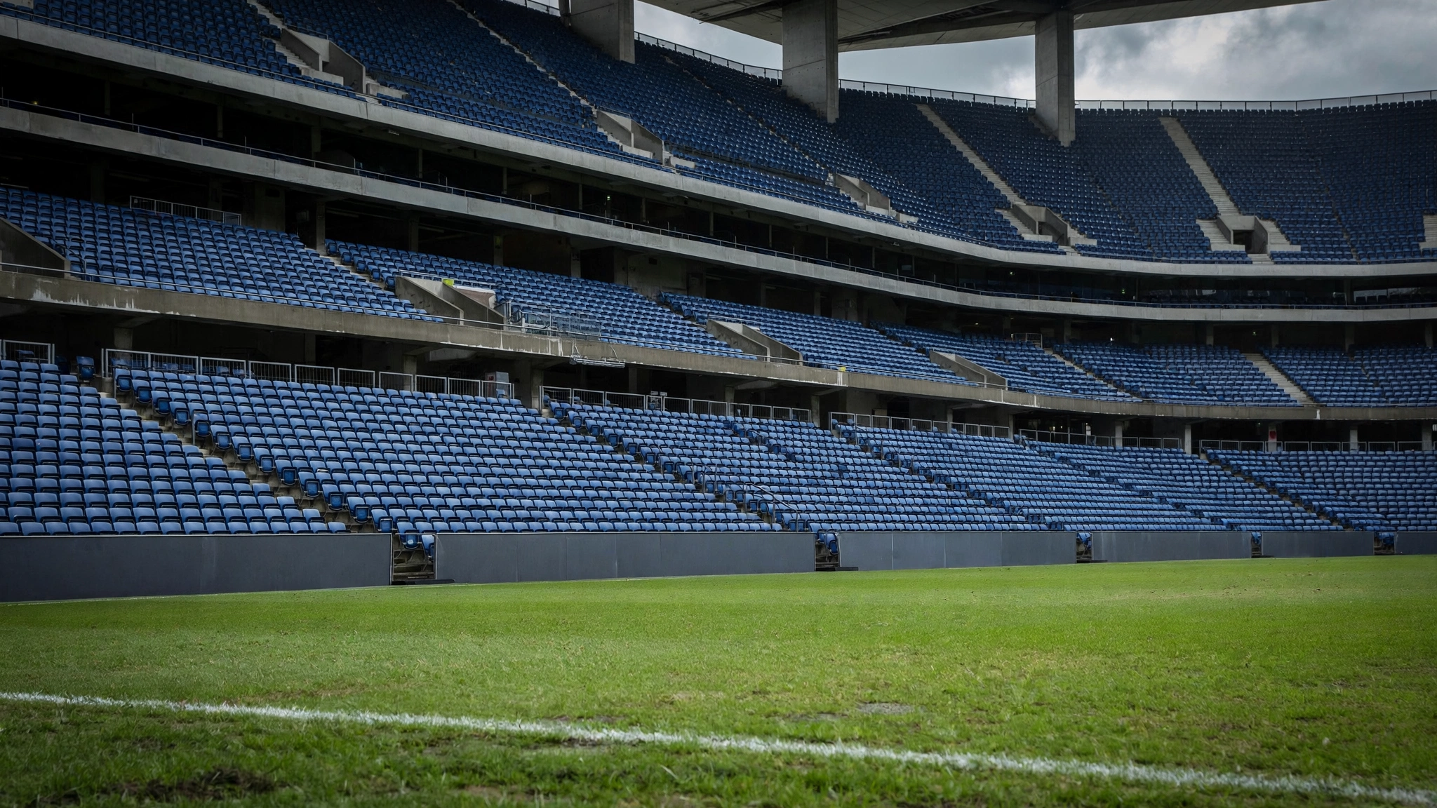 Rows of empty blue seats in a large soccer stadium above a grass pitch