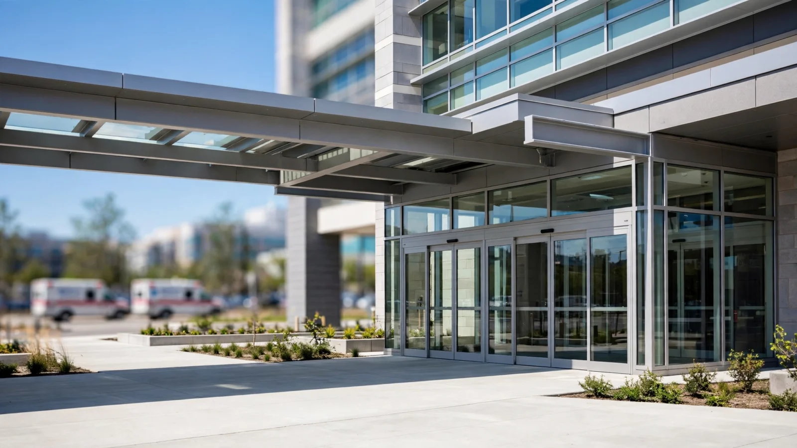 Modern hospital entrance with glass doors under a canopy on a clear day, with ambulances parked in the blurred background