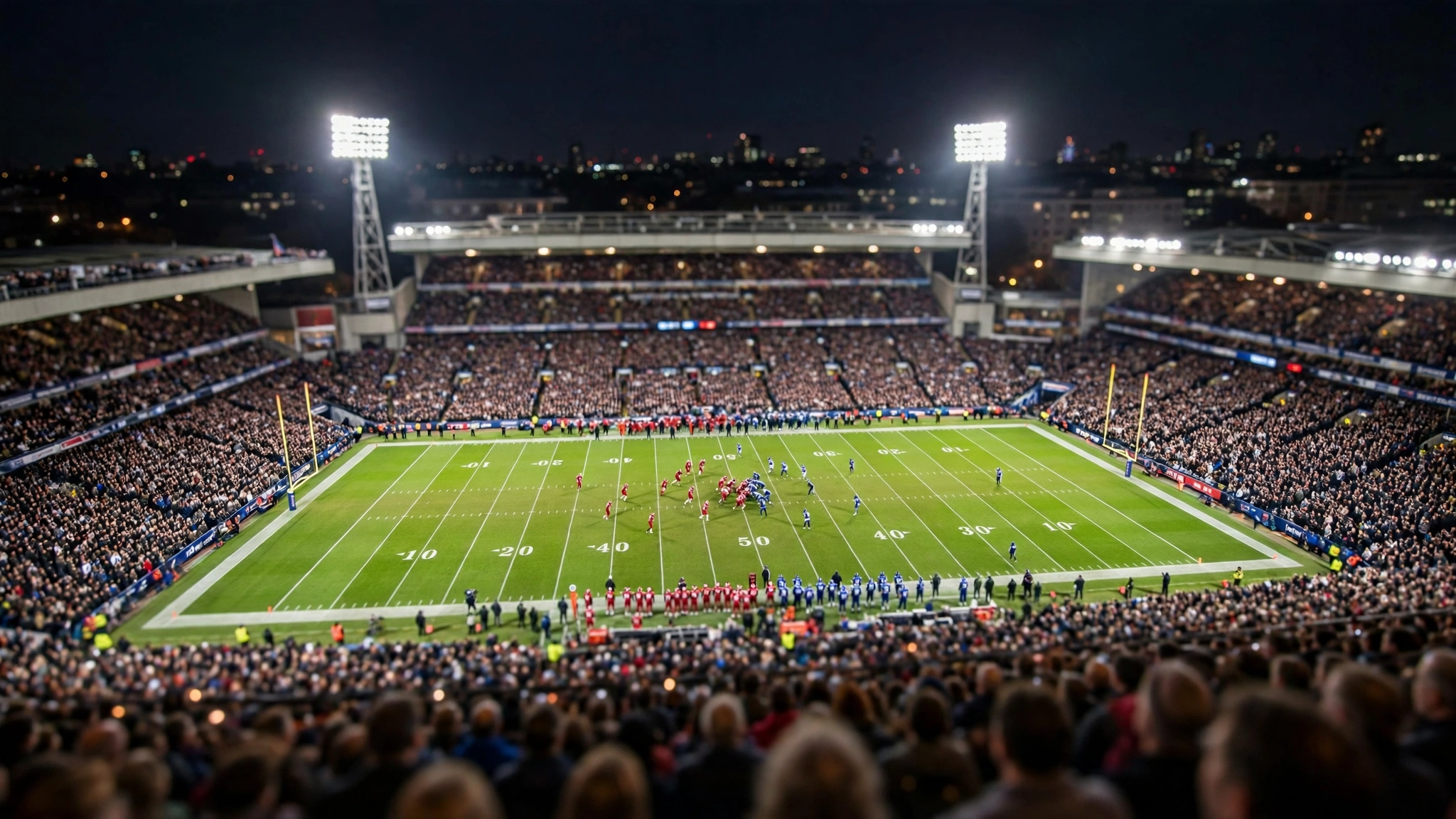 Packed American football stadium at night with a green field lit by floodlights and players in red and blue uniforms