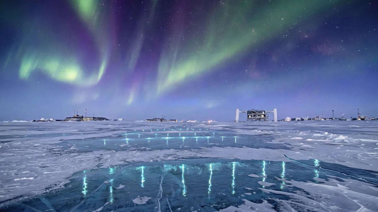 Antarctic ice plain at night under aurora with research station buildings in the distance and faint lights glowing beneath the ice