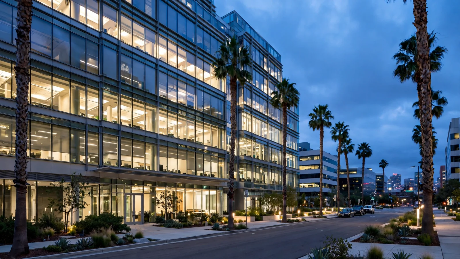 A modern glass office building with lit windows at dusk, lined with palm trees along the sidewalk
