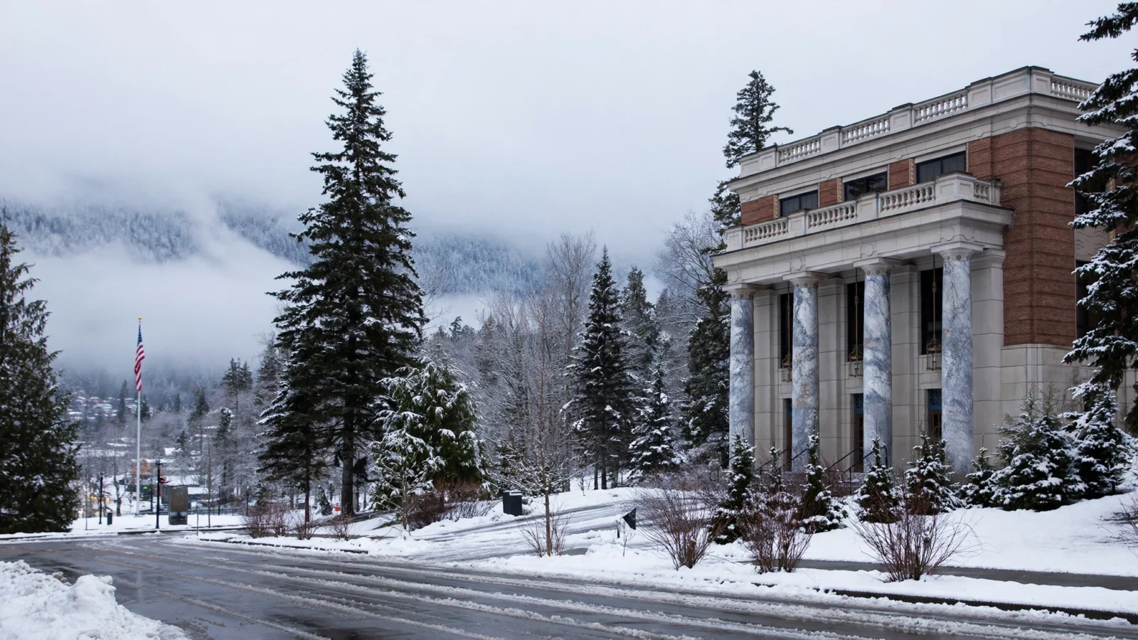A state capitol building with marble columns in a snowy Alaskan town, with an American flag and snow-covered evergreen trees