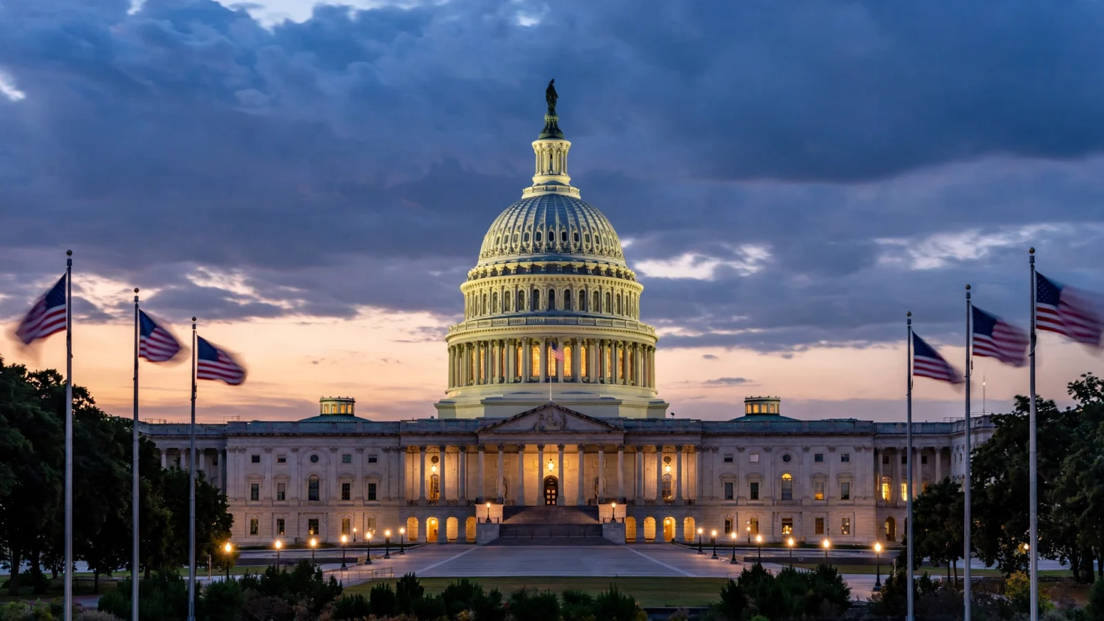 The U.S. Capitol dome at dusk with American flags flying in front of the building.