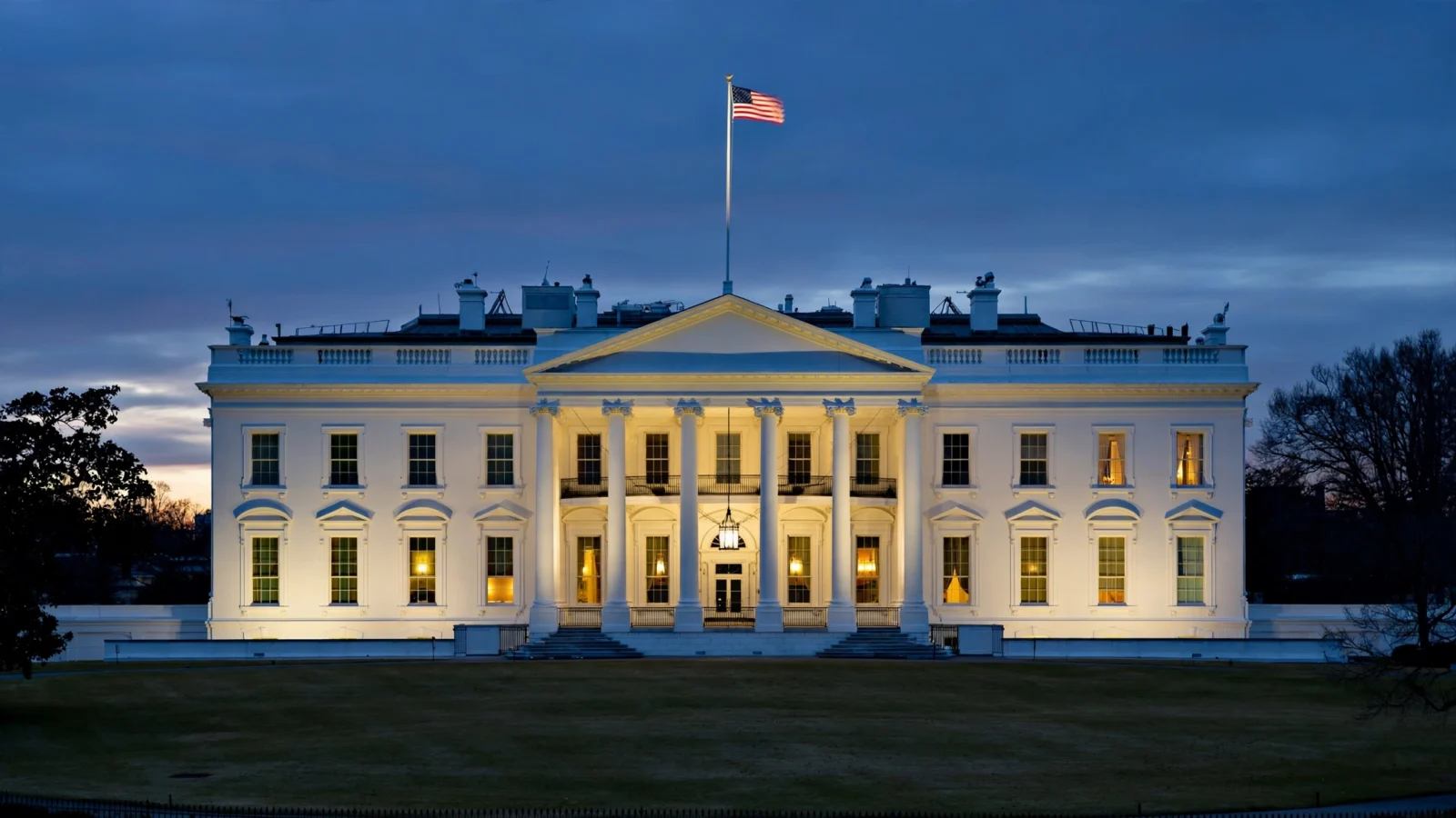 The White House north facade glowing at dusk, the American flag flying from the rooftop flagpole above the columned portico, with an empty lawn in the foreground.
