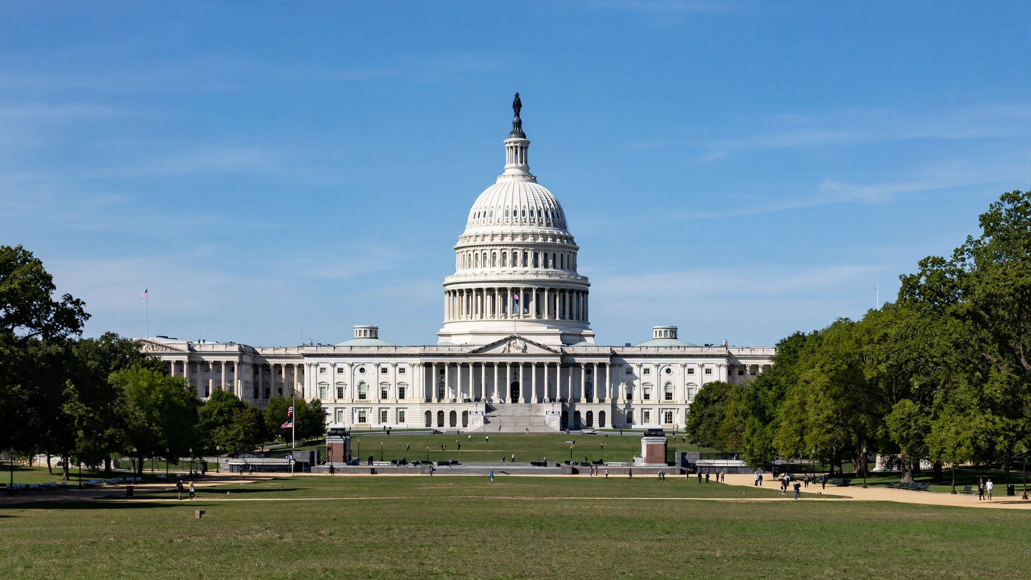 A wide view of the U.S. Capitol building with its white dome under a clear blue sky, framed by green trees and the lawn in front.