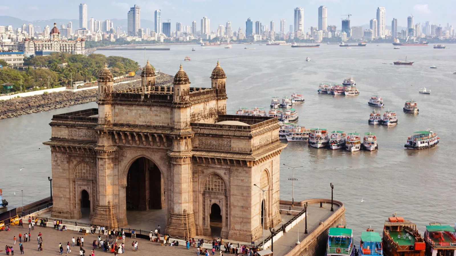 Aerial view of the Gateway of India monument on the Mumbai waterfront, with boats in the harbor and the city skyline behind
