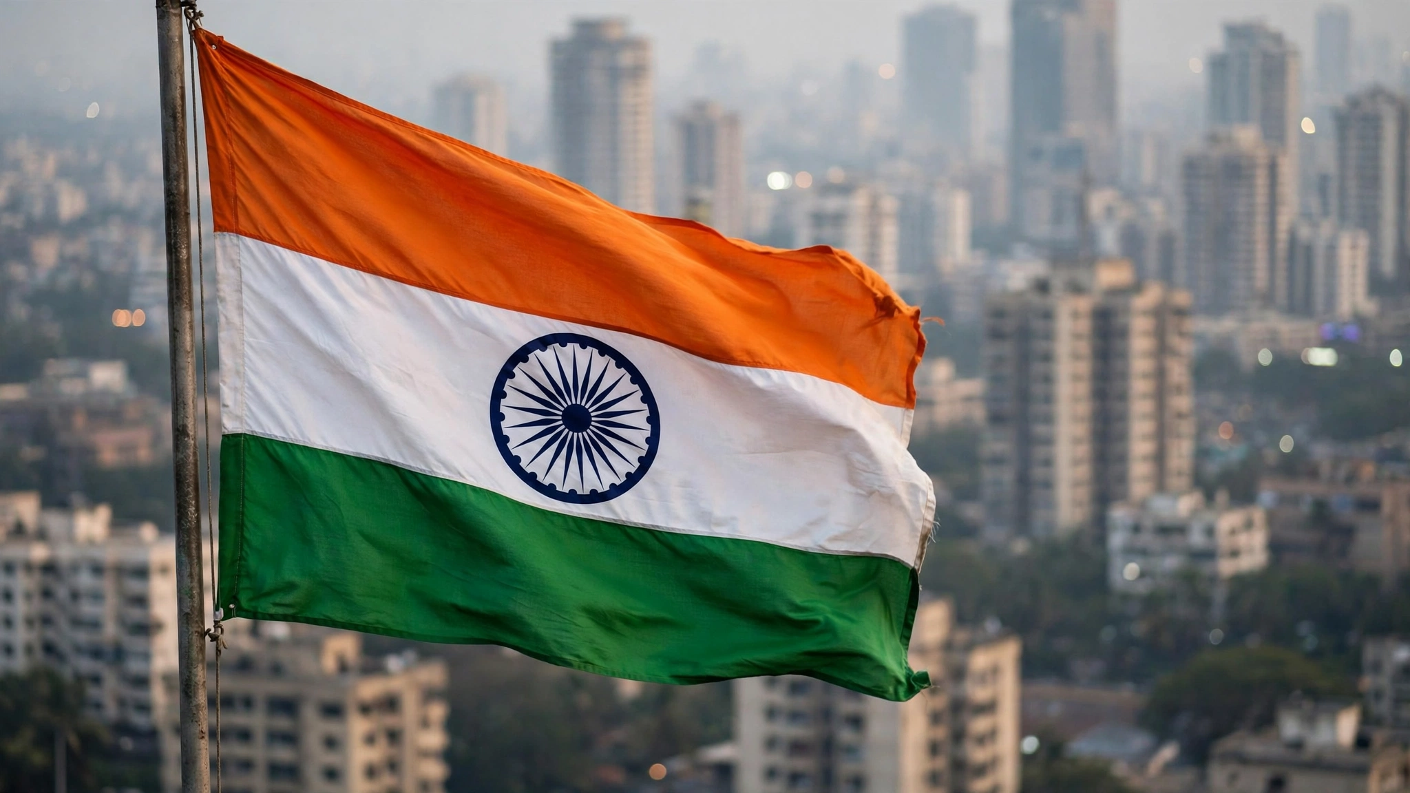 The Indian national flag waving on a pole with blurred city buildings in the background