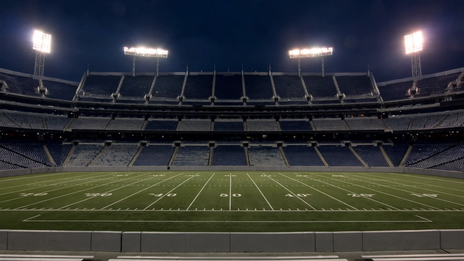 An empty football stadium at night with floodlights on over a green field marked with yard lines.