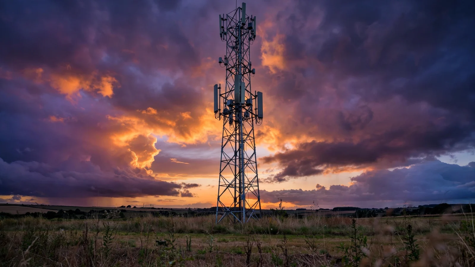 A cell tower silhouetted against an orange and purple dusk sky, with a rural field in the foreground