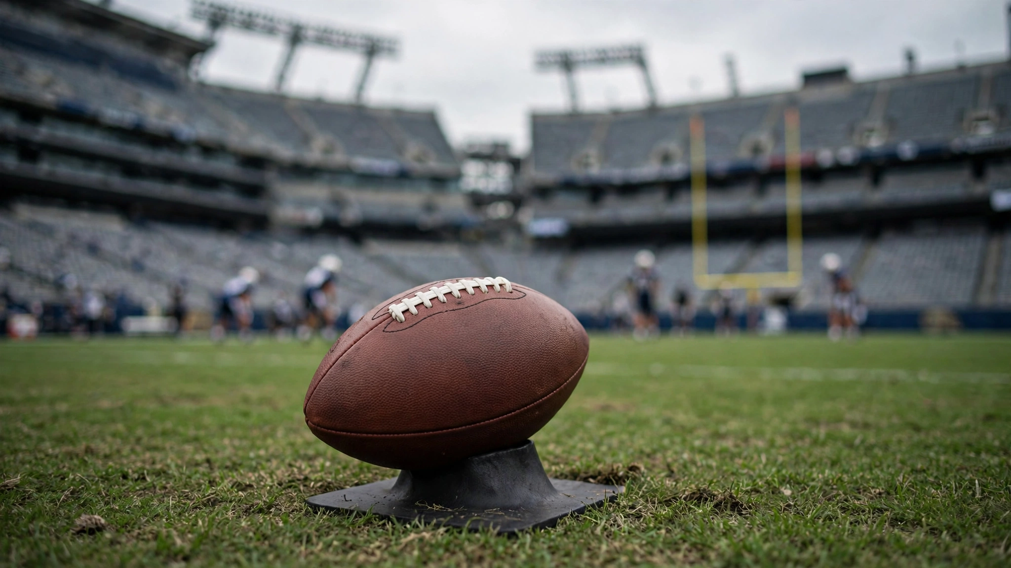 An American football on a kicking tee on a grass field with goalposts and players blurred in the background.