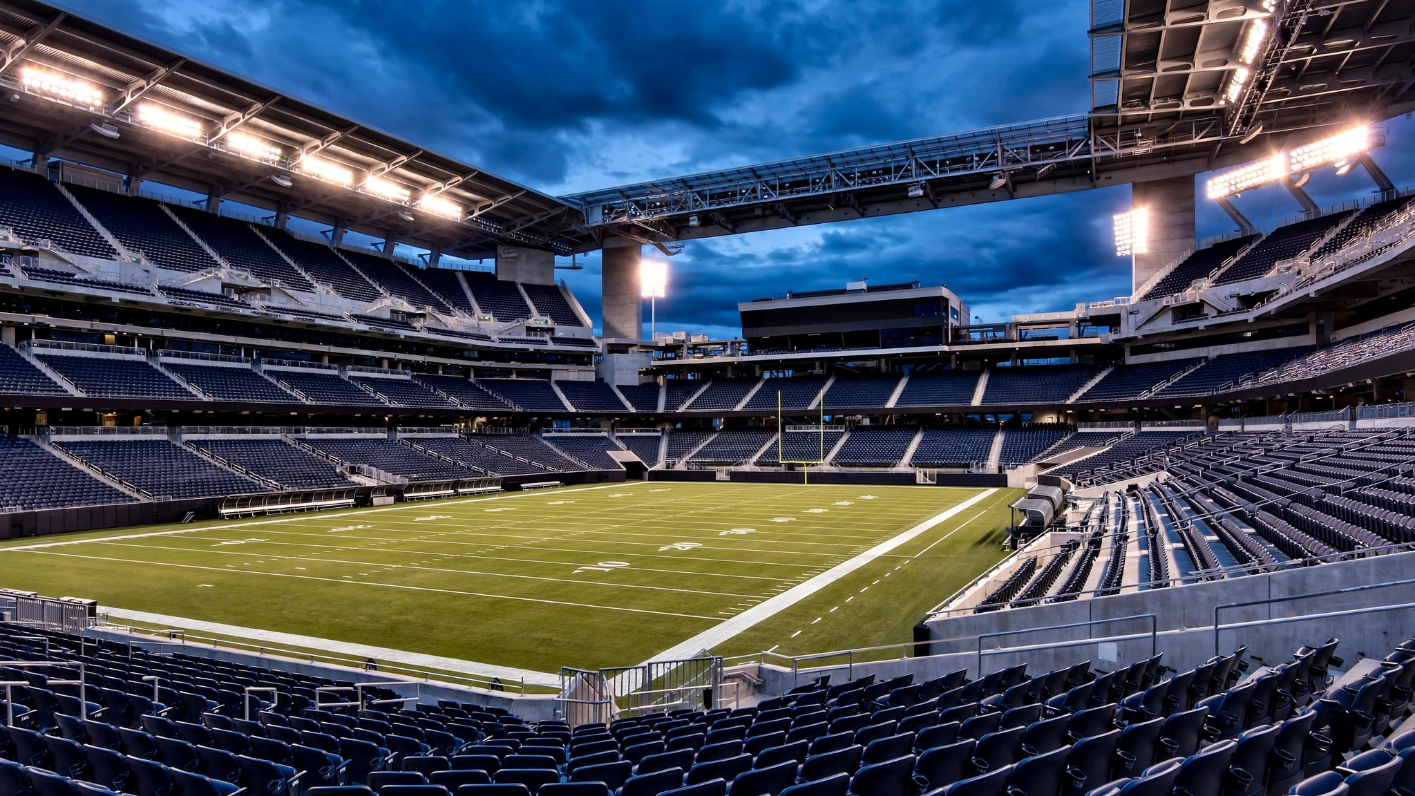 An empty modern football stadium interior at dusk, with a lit green field, tiered empty stands and glowing floodlights under a dark cloudy sky.