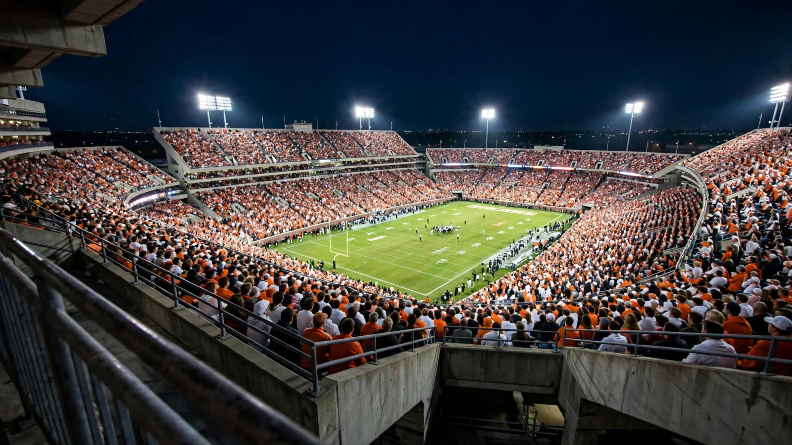 A packed college football stadium in orange and white under floodlights at night.