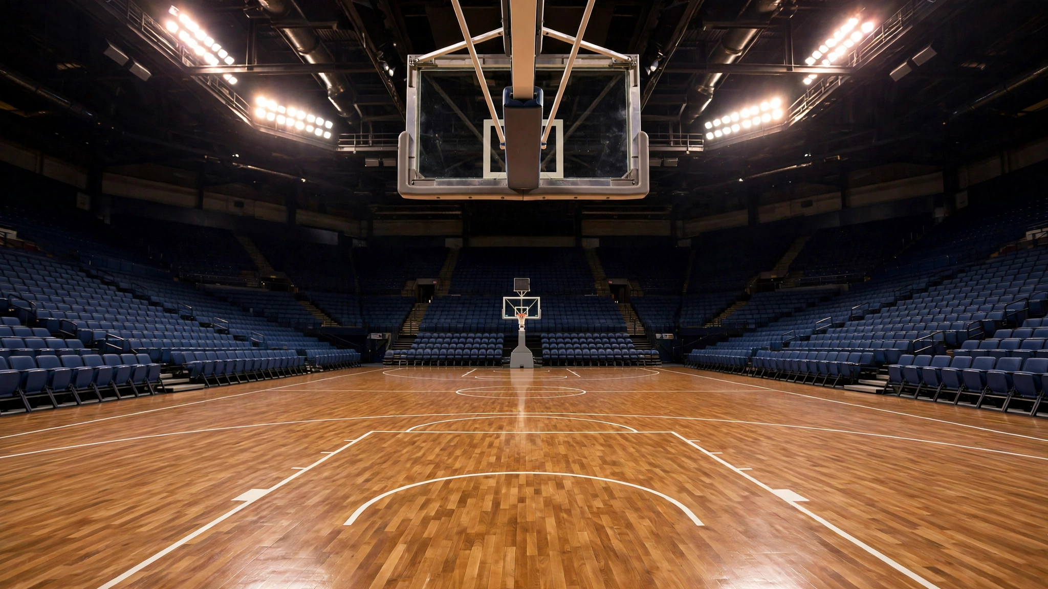 An empty basketball court with a polished wooden floor and hoops under bright arena lights, with empty blue seats around it