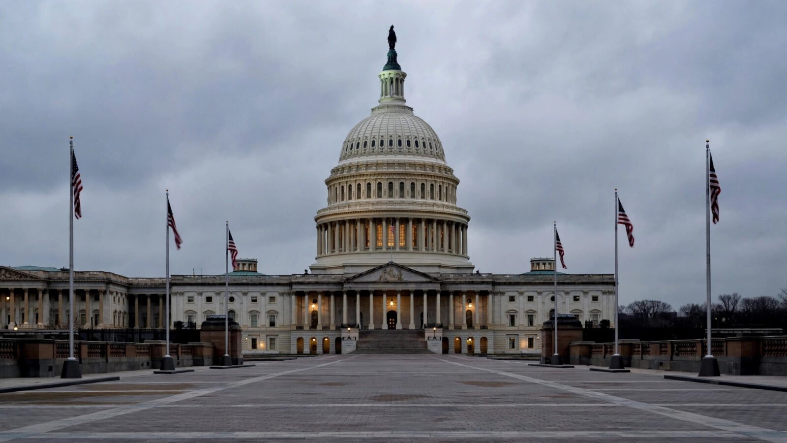 The U.S. Capitol dome at dusk, viewed across an empty plaza, with American flags on flagpoles.