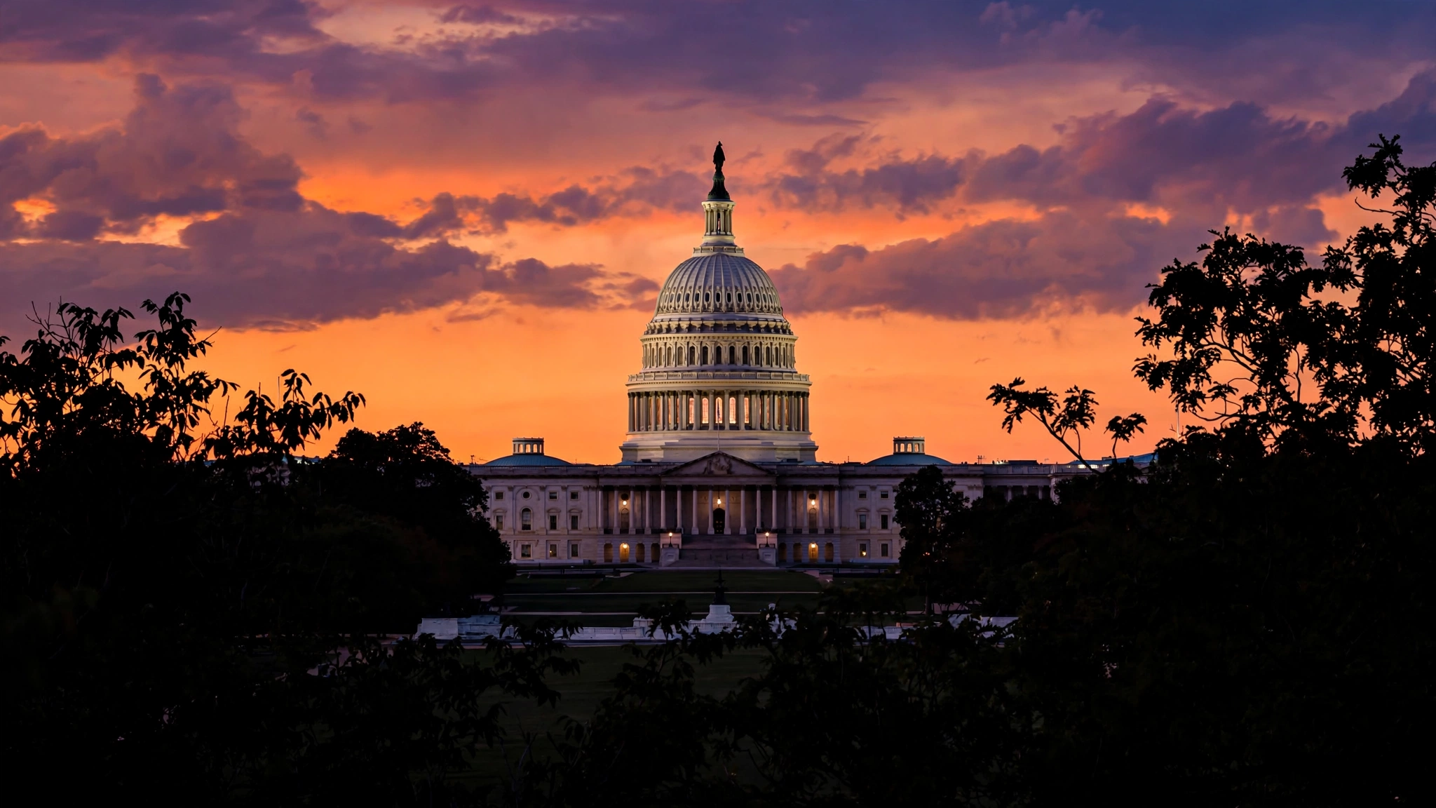 The US Capitol dome at sunset under an orange and purple sky, framed by silhouetted trees.