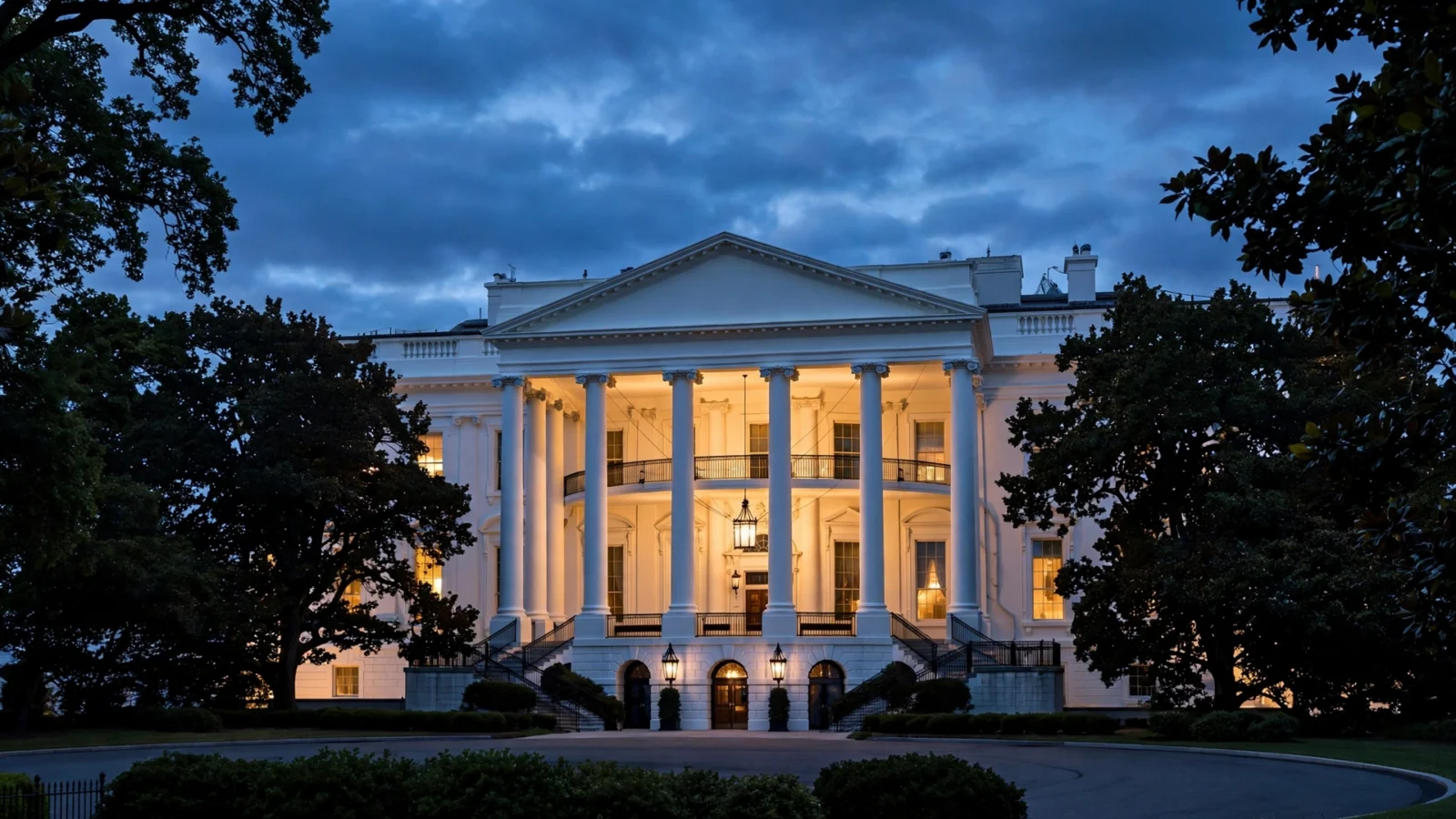 The White House North Portico with its windows lit at dusk, framed by trees under a cloudy evening sky.