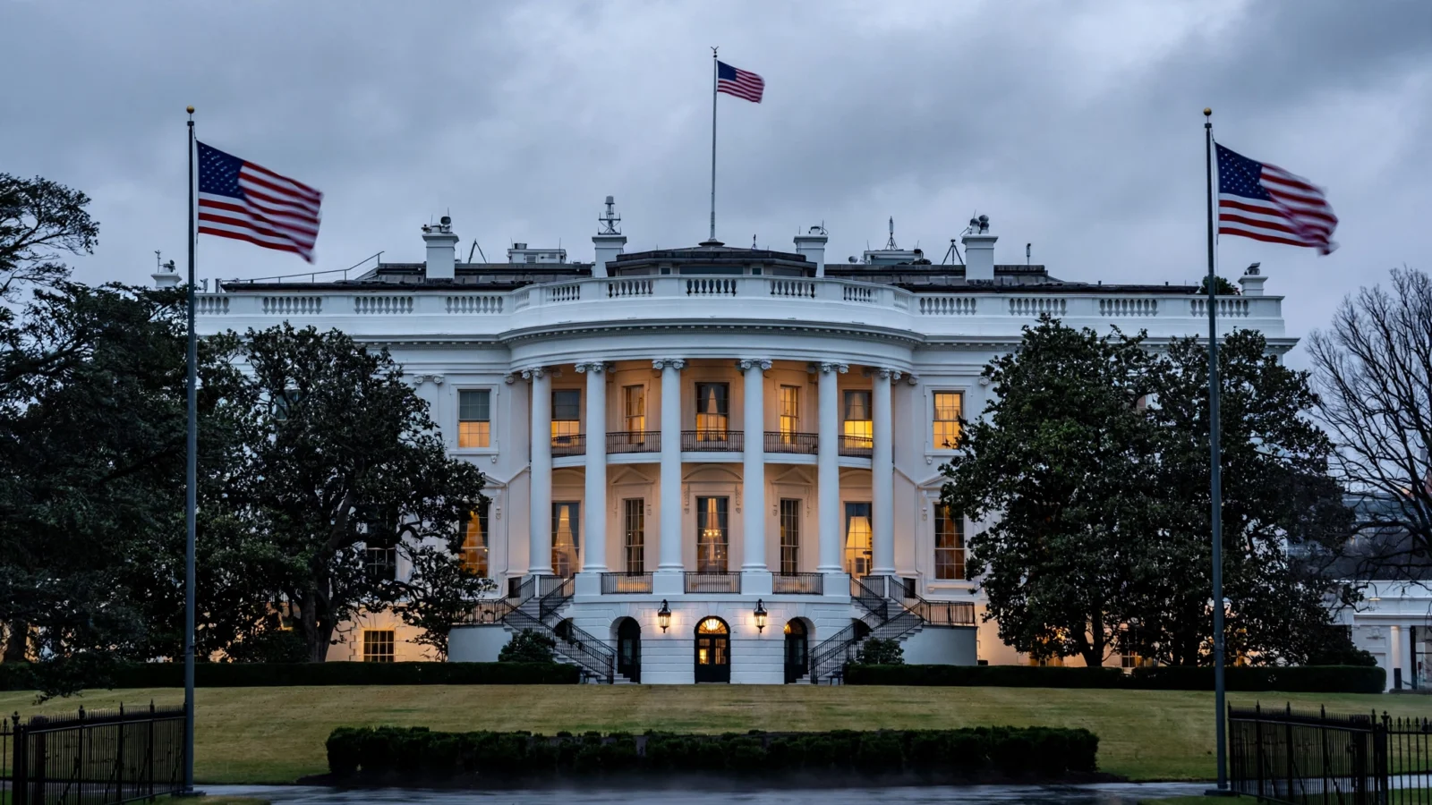 The White House South Portico at dusk, with American flags flying under an overcast sky.