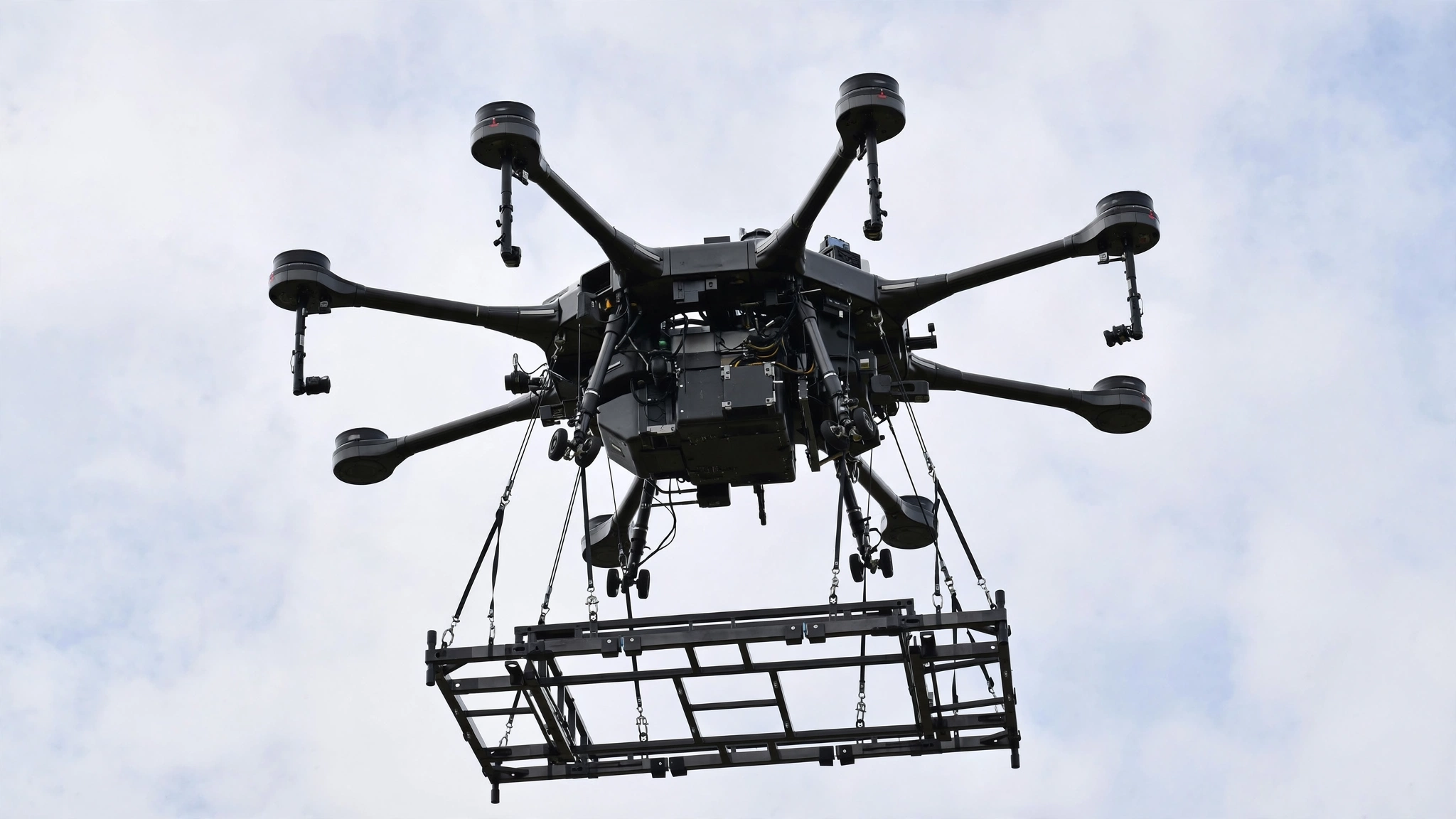 A heavy-lift hexacopter drone seen from below against a cloudy sky, carrying a suspended transport frame.