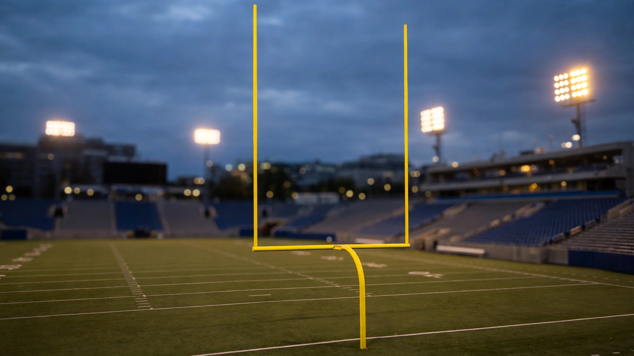 Yellow goal posts stand on a football field at dusk with stadium lights on