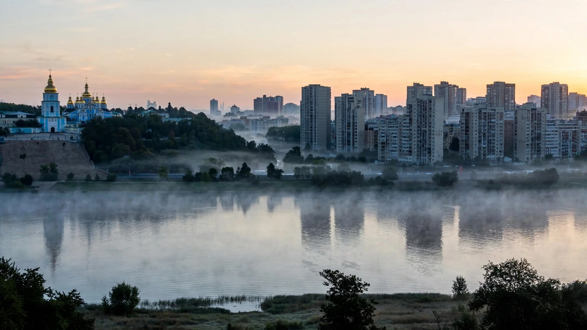 Residential towers and a golden-domed church across the Dnipro River at dawn
