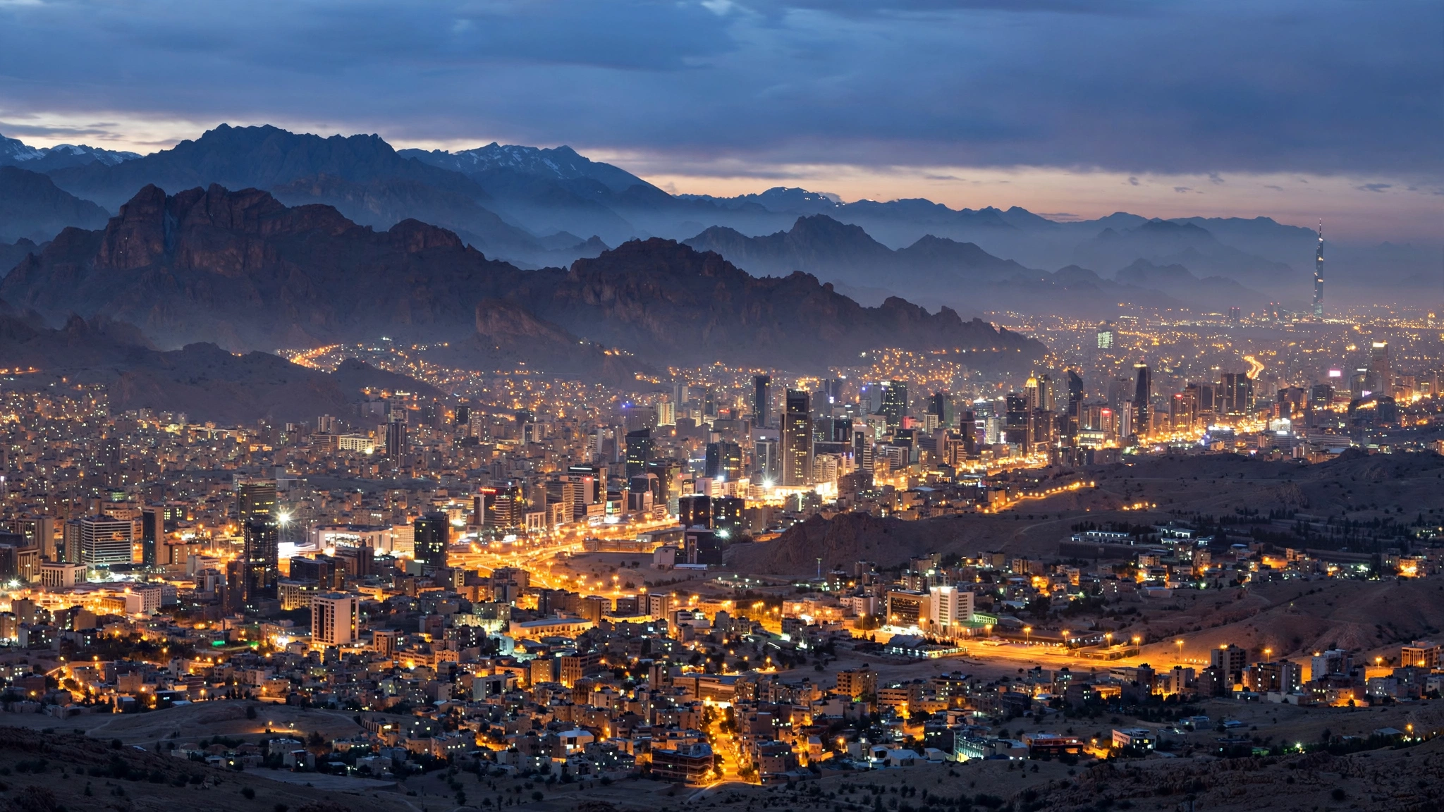 A Middle Eastern capital city skyline at dusk with mountains behind it