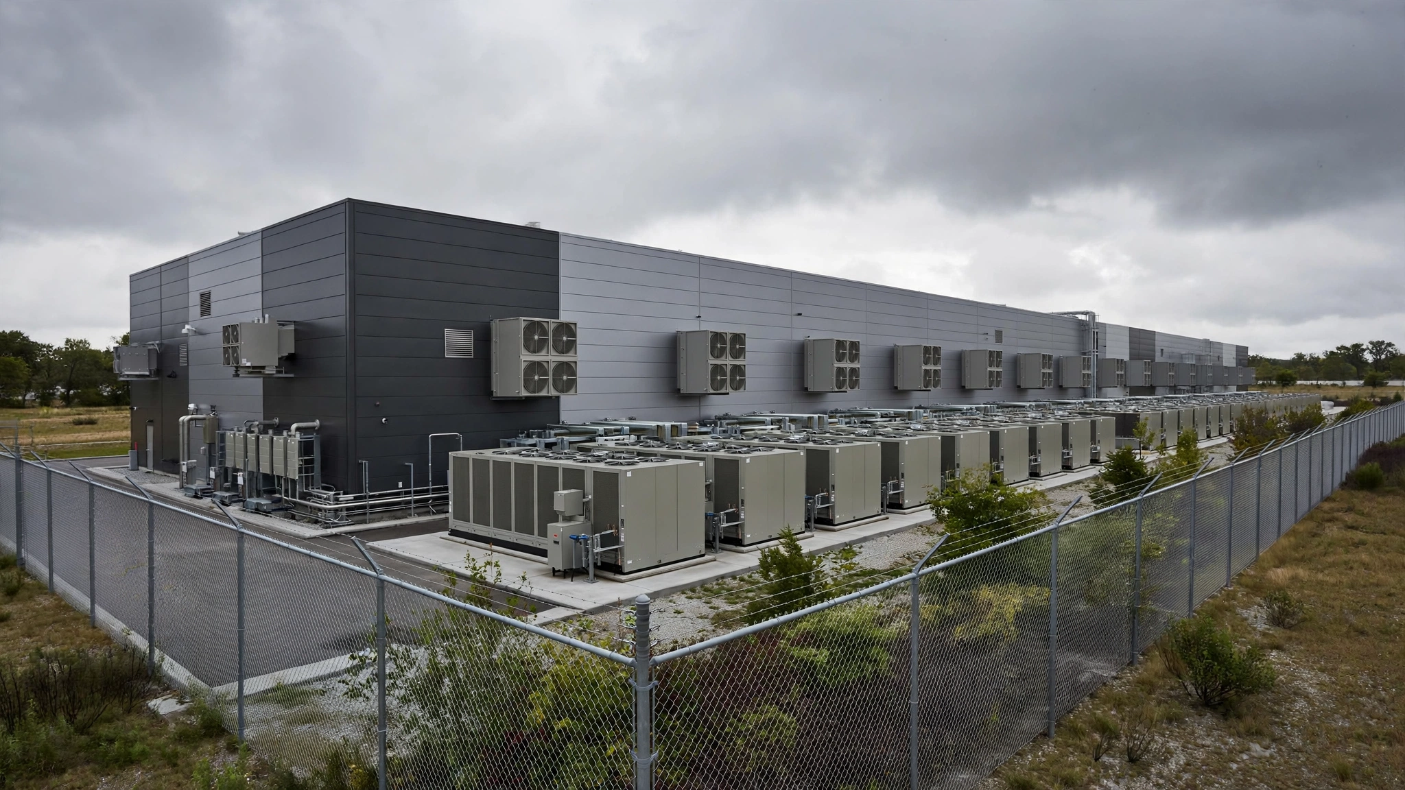 A large data center building with rows of outdoor cooling units behind a fence