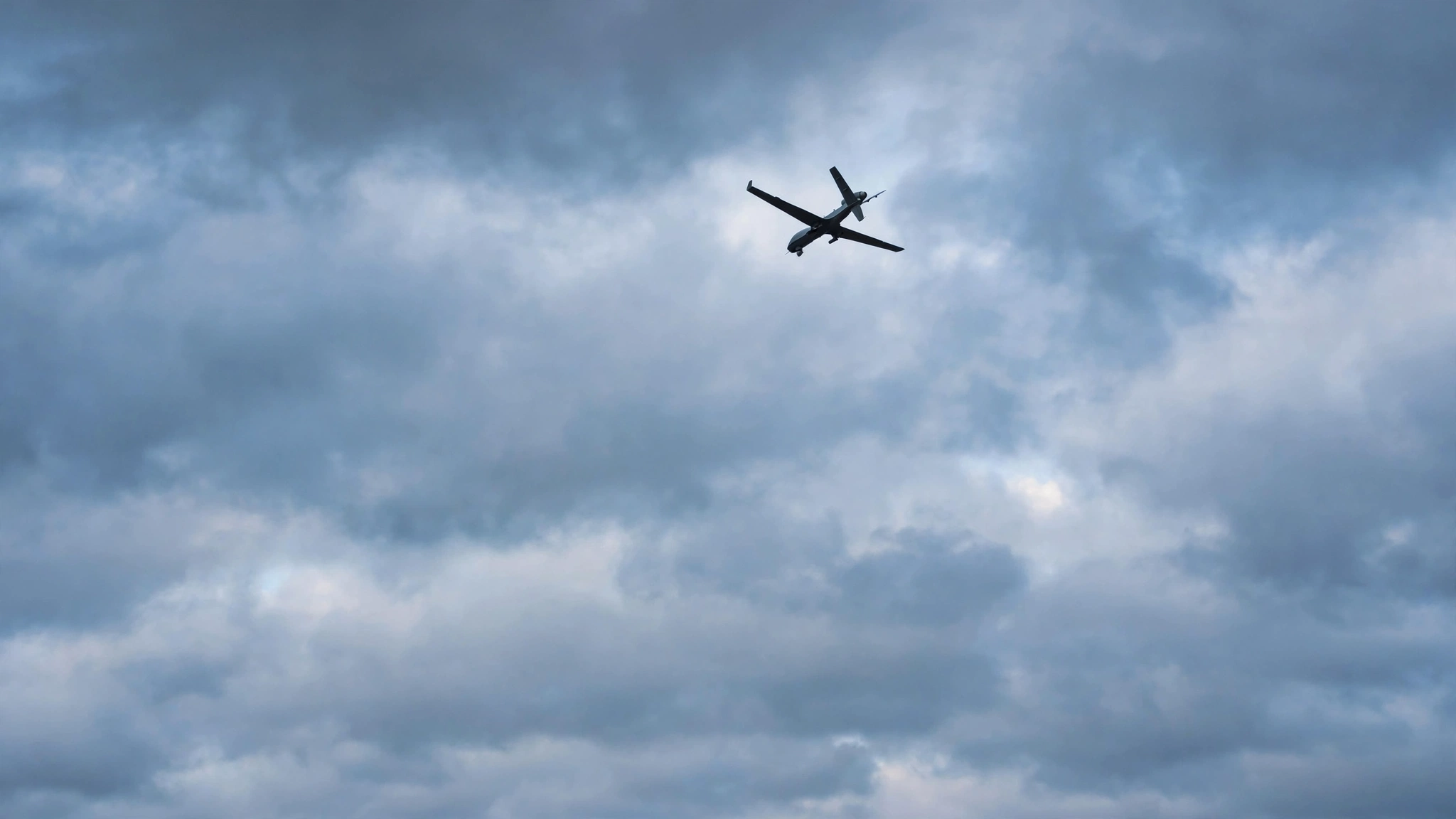 A gray military-style drone flying high in a cloudy blue-gray sky