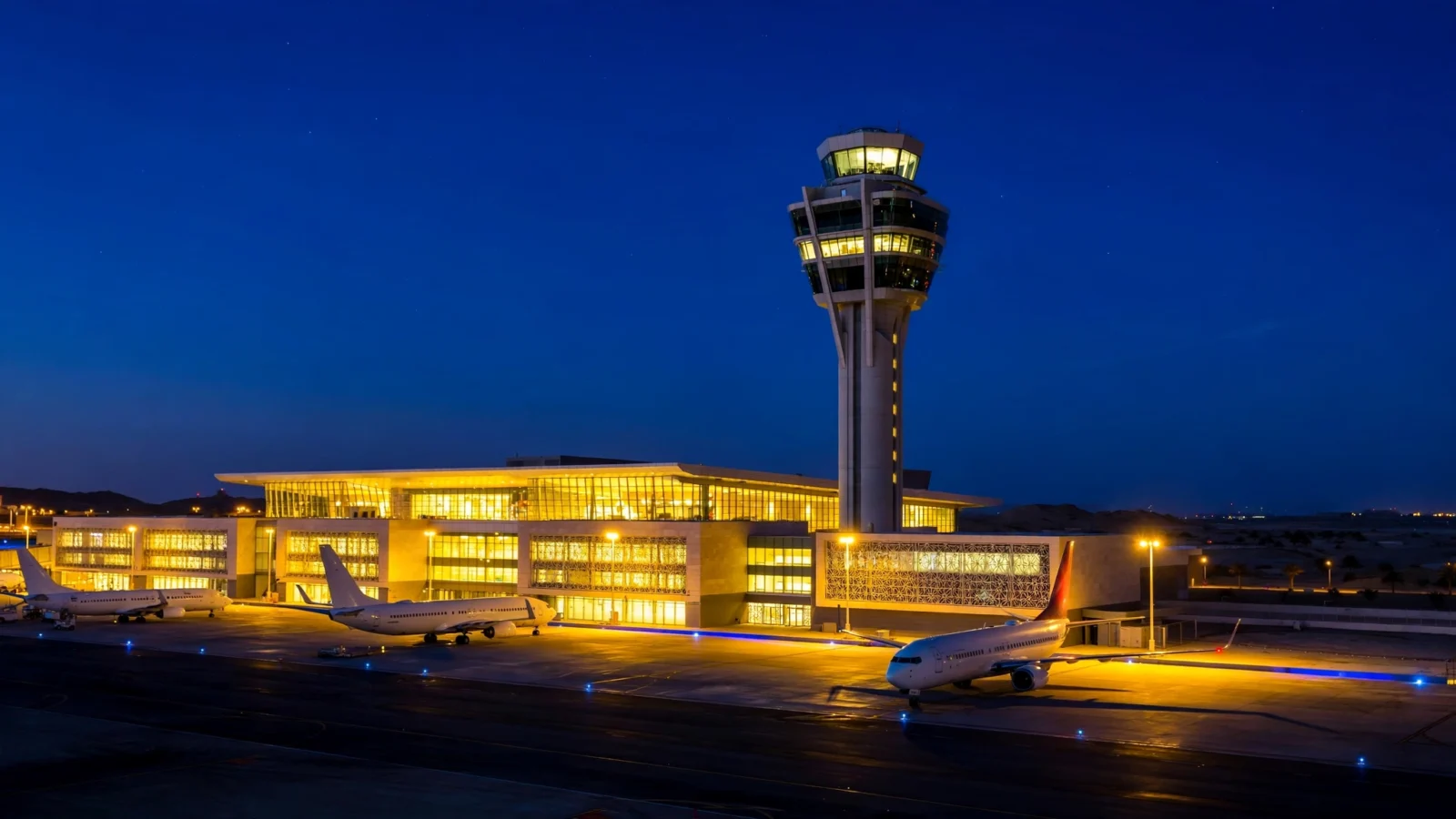 A lit control tower and airport terminal at night with passenger aircraft parked on the tarmac, no visible signs or logos.