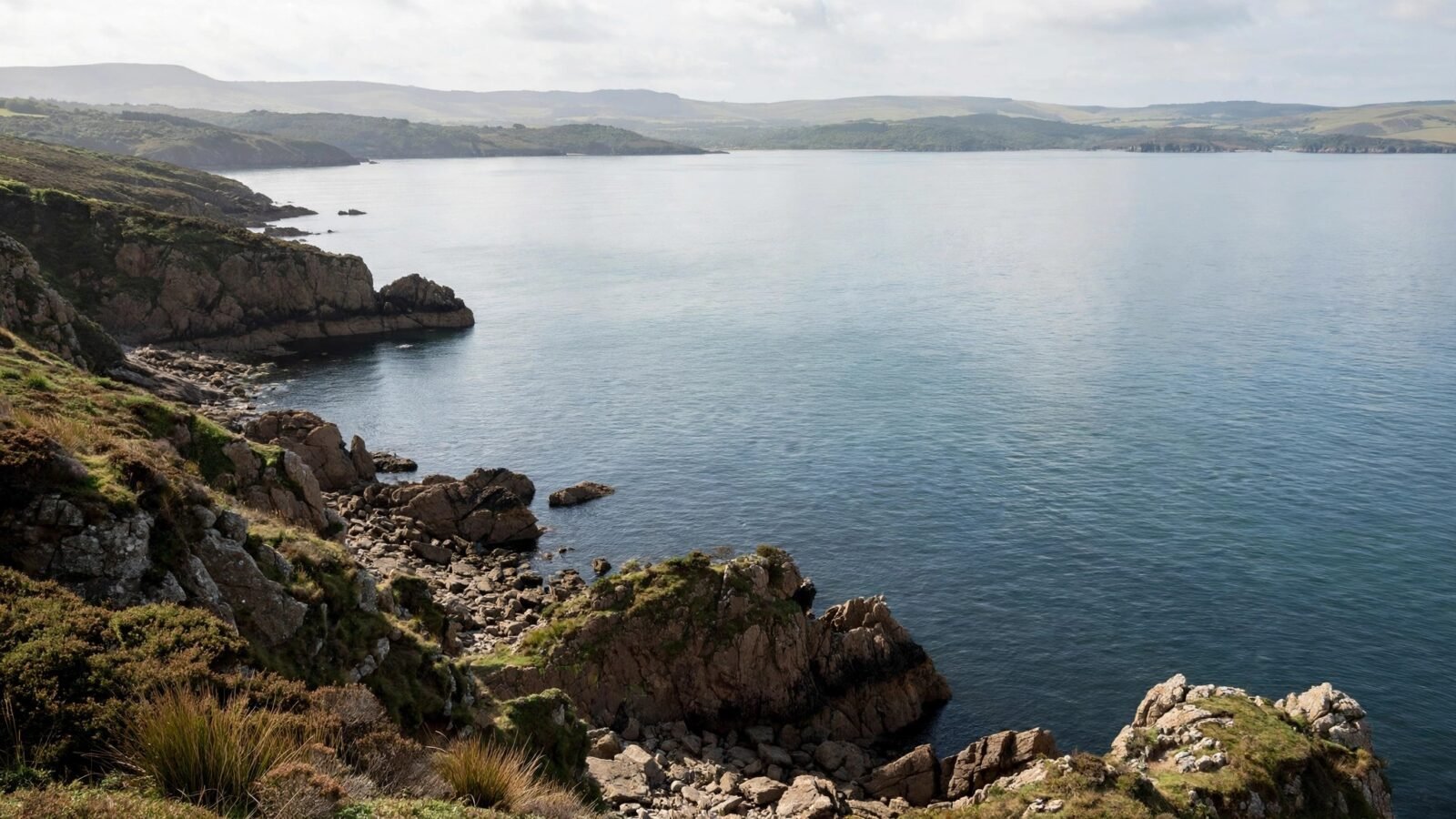 A rocky coastline with calm sea water and green hills in the distance under a cloudy sky