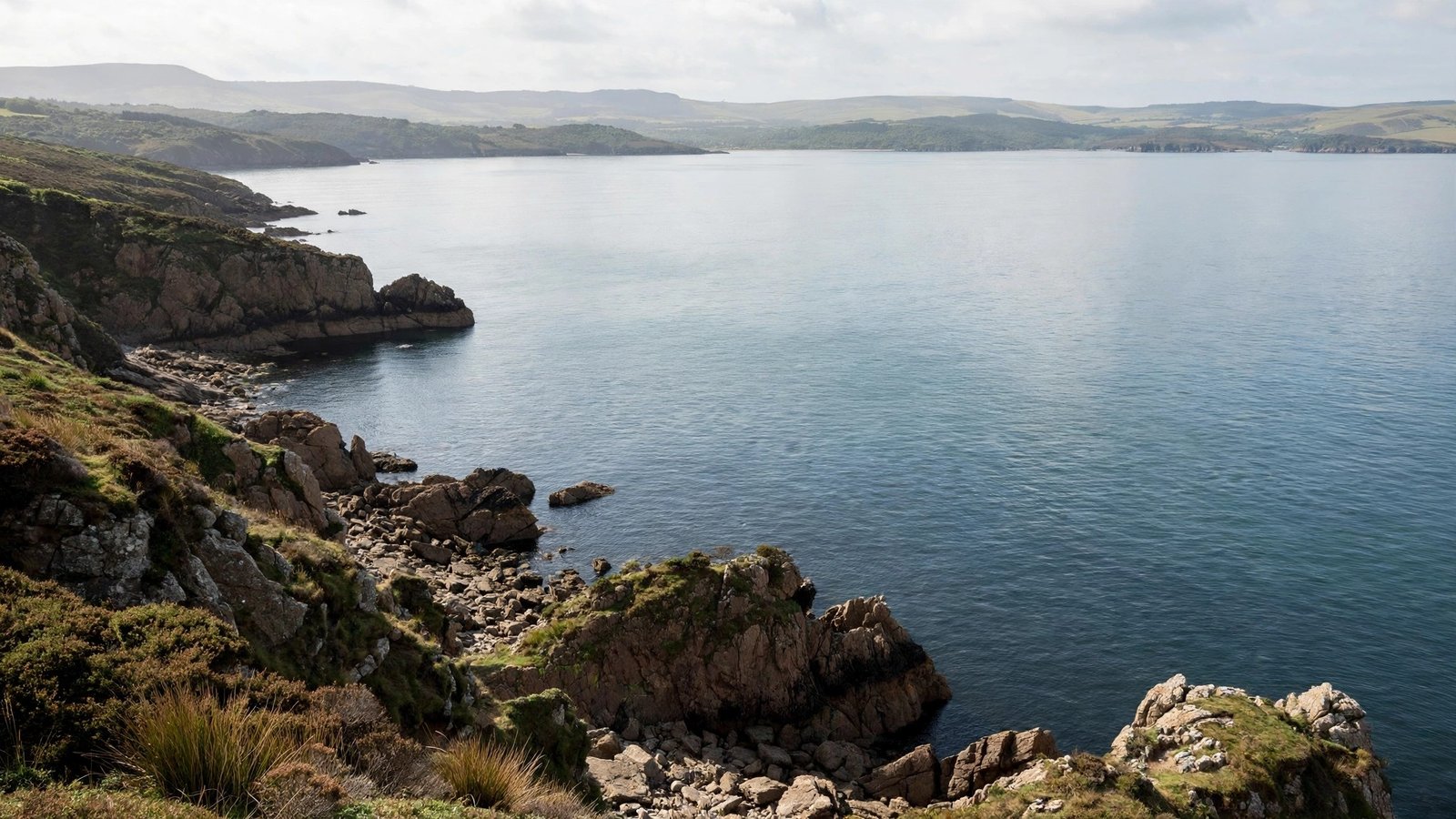 A rocky coastline with calm sea water and green hills in the distance under a cloudy sky