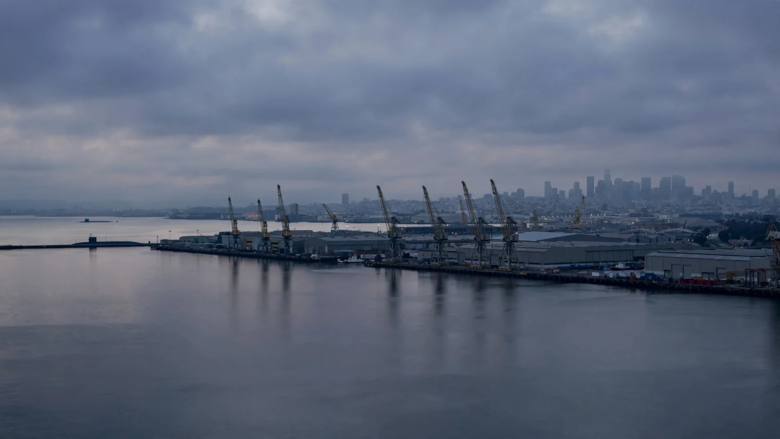 An industrial harbor shipyard with cranes and warehouse buildings along calm water at dusk, a submarine visible in the distance and a city skyline behind.