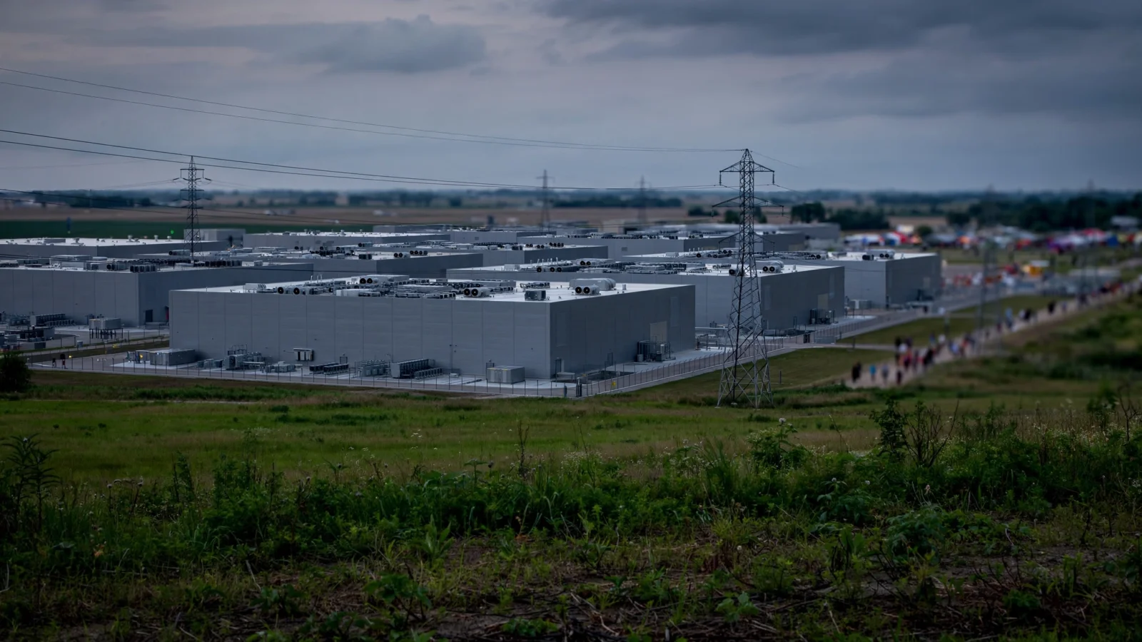 A large gray data center campus with rooftop cooling equipment, power transmission towers and green fields in the foreground under a cloudy sky
