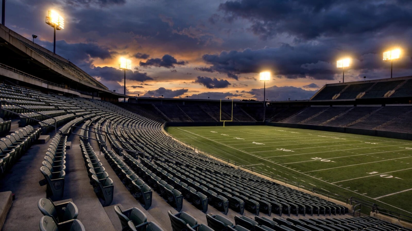 An empty American football stadium at dusk with the field lit by floodlights.