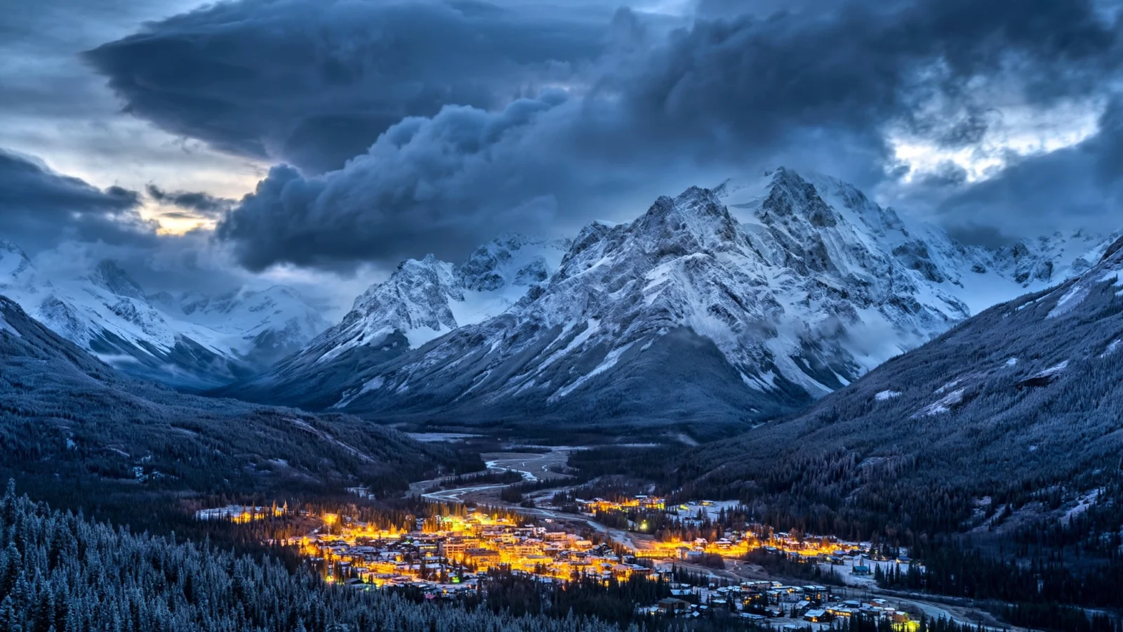 Snow-capped Alaska mountain peaks at dusk with town lights glowing in the valley below